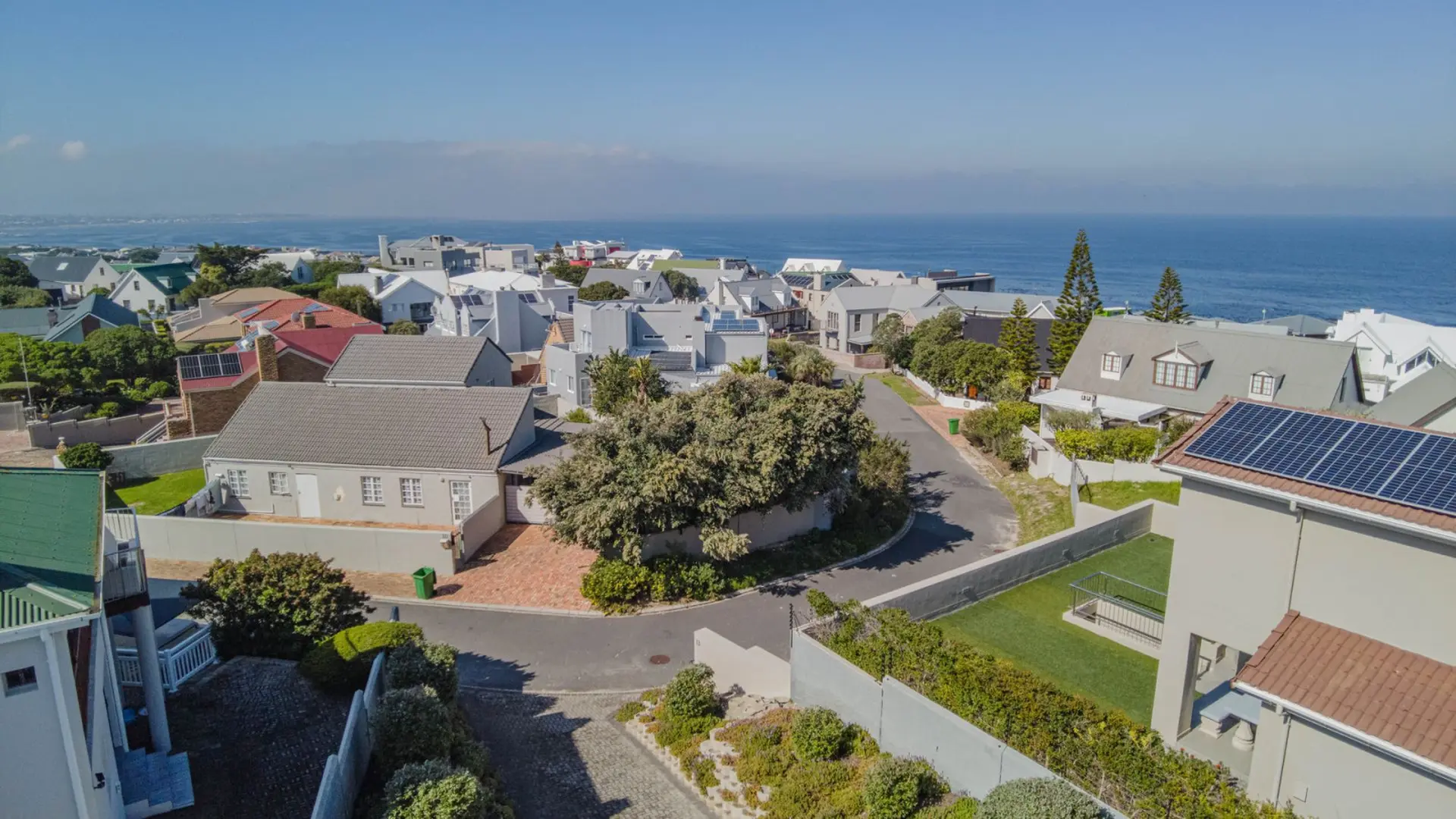Aerial view of a coastal neighborhood with houses, trees, and the ocean in the background.