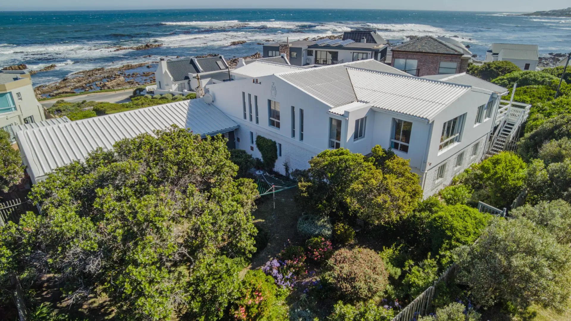 An aerial view of a white house with a white metal roof and a lush green garden, overlooking the oce