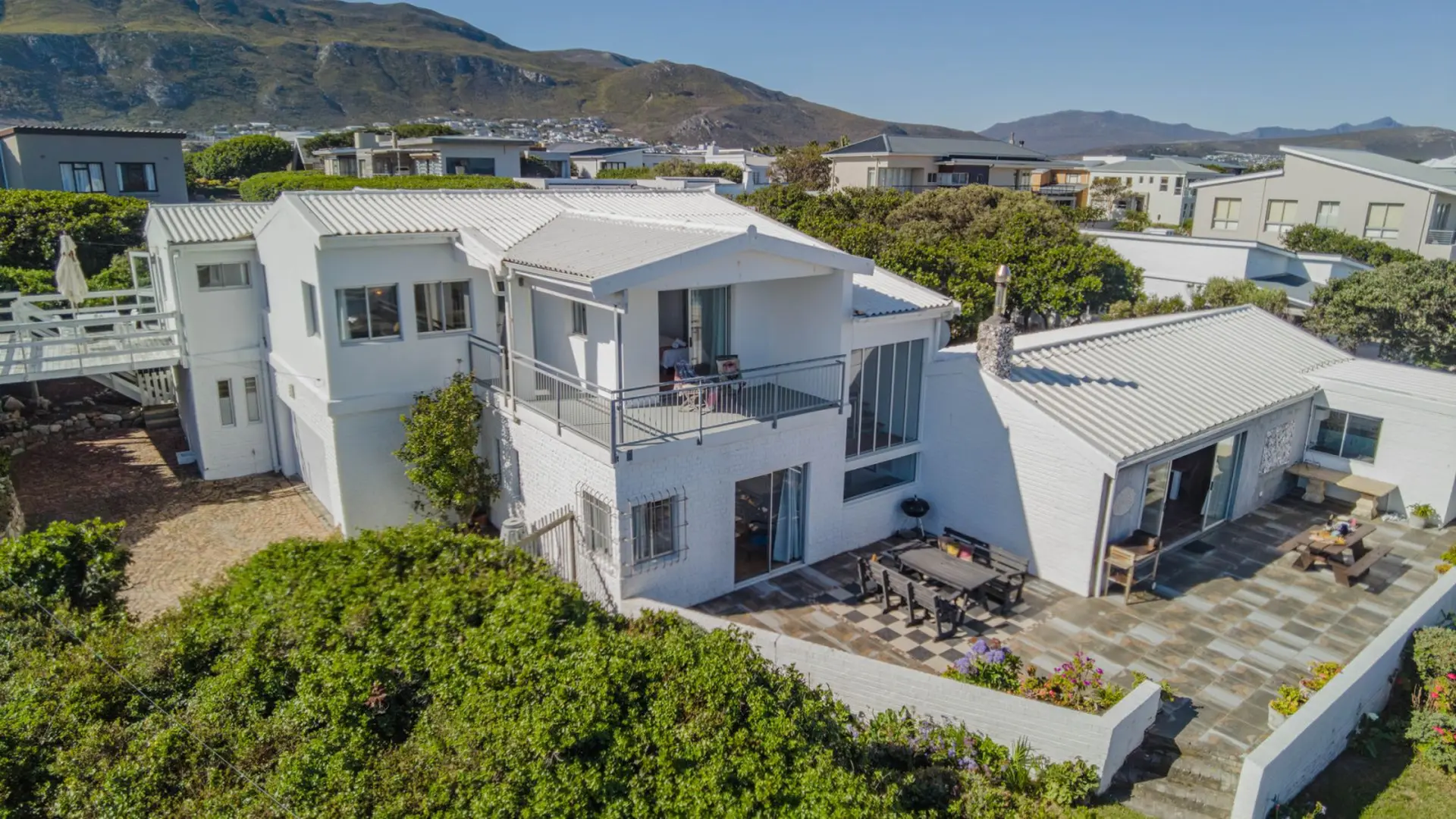 A drone shot of a modern white house with an upstairs balcony, patio, and garden, overlooking mounta