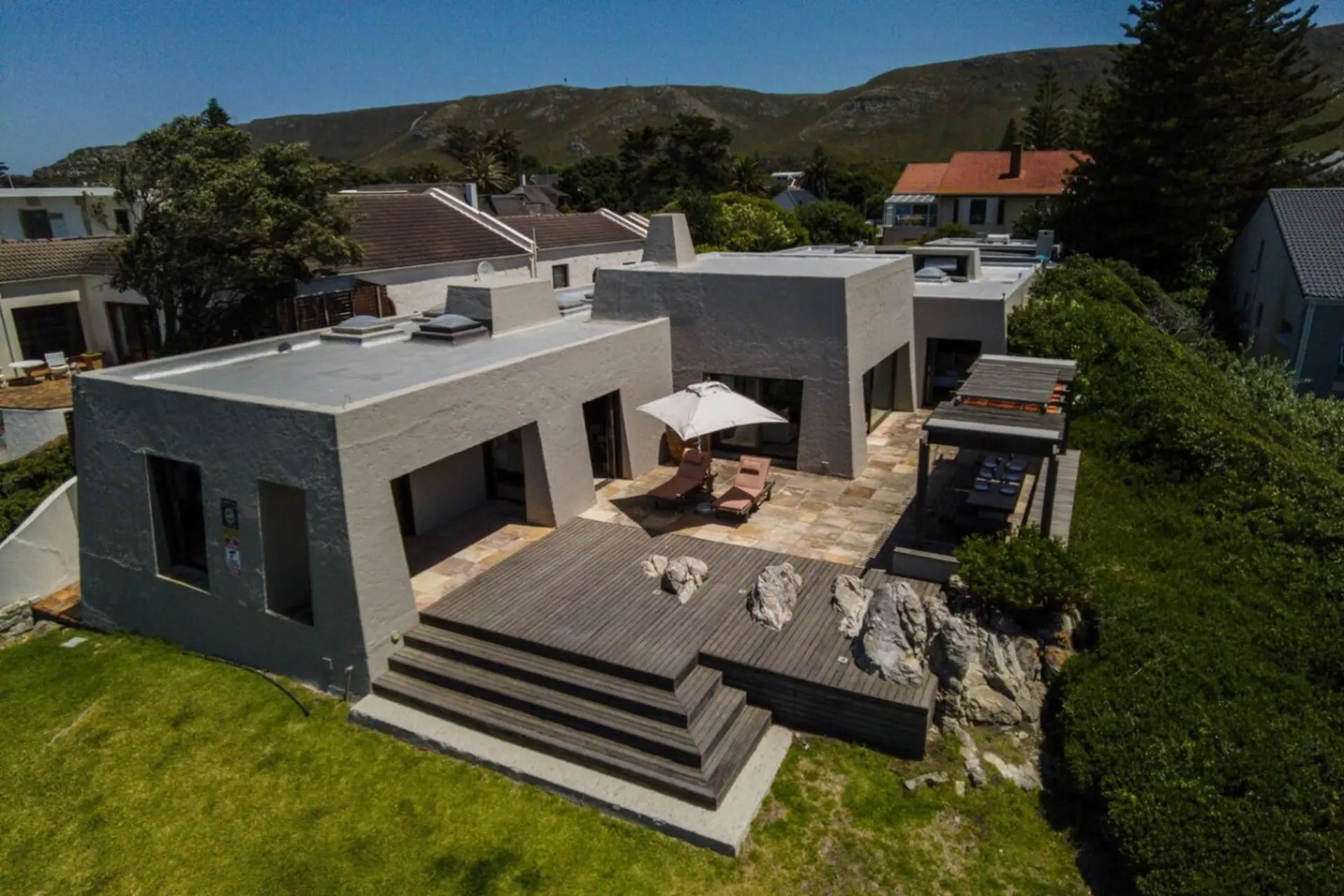 A modern house with a large deck and outdoor seating area sits on a grassy hill.