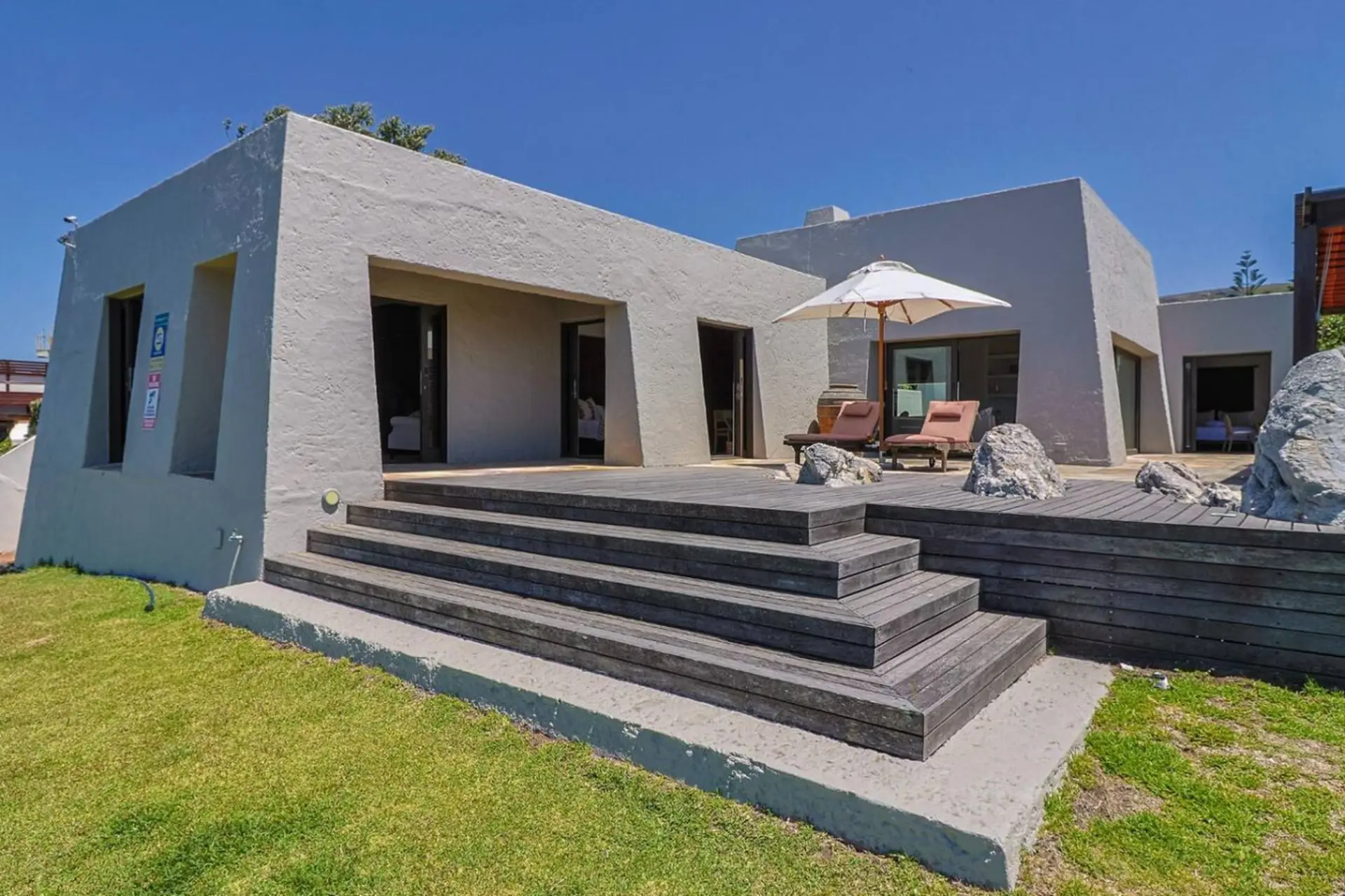 Modern gray house with a wooden deck and lounge chairs, under a clear blue sky.