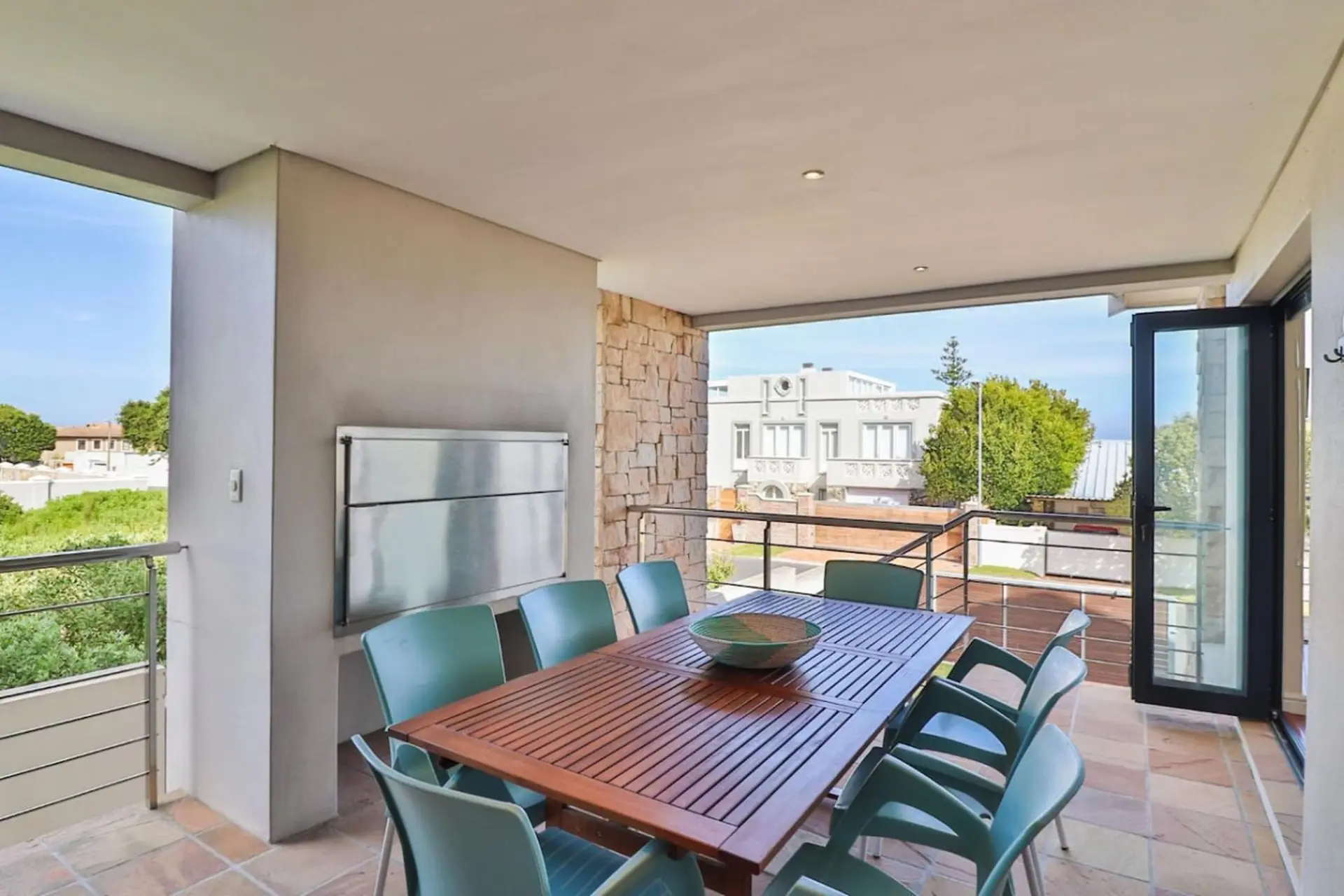 A patio with a table, chairs, and grill overlooks houses and trees.