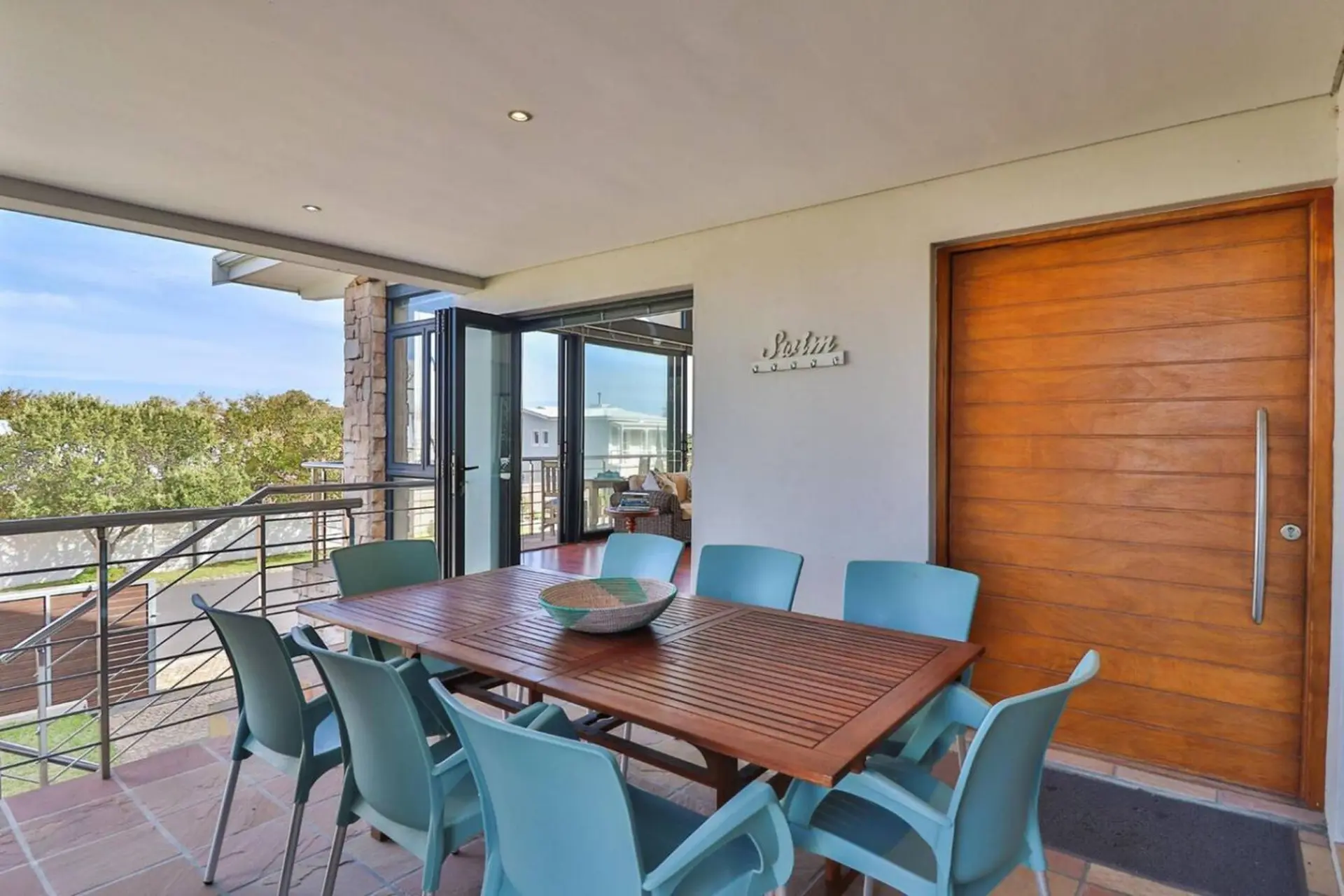 Outdoor dining area with wooden table and chairs, facing a balcony and trees.