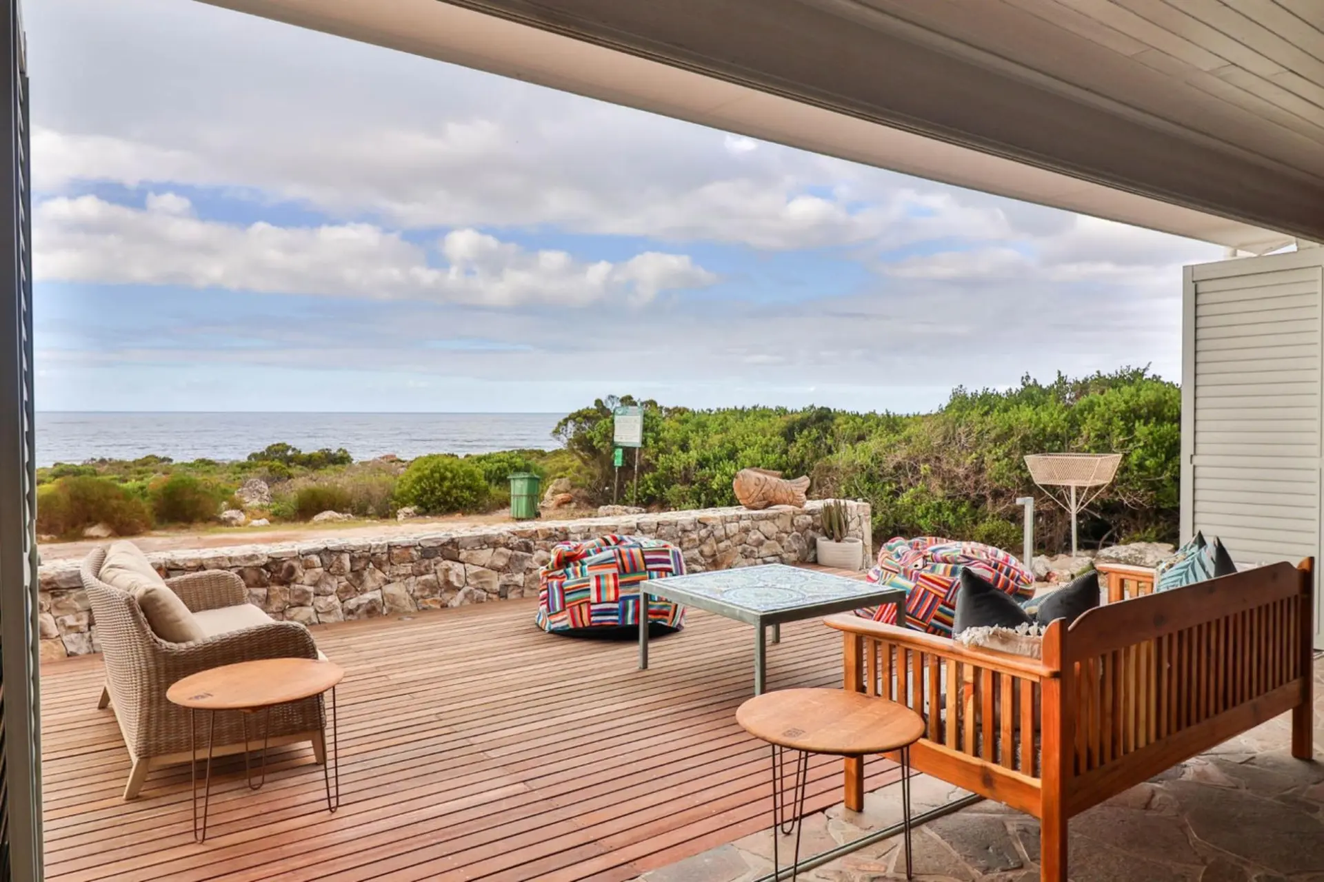 Outdoor patio with wooden deck overlooking the ocean, furnished with wicker sofa, beanbags, and wood