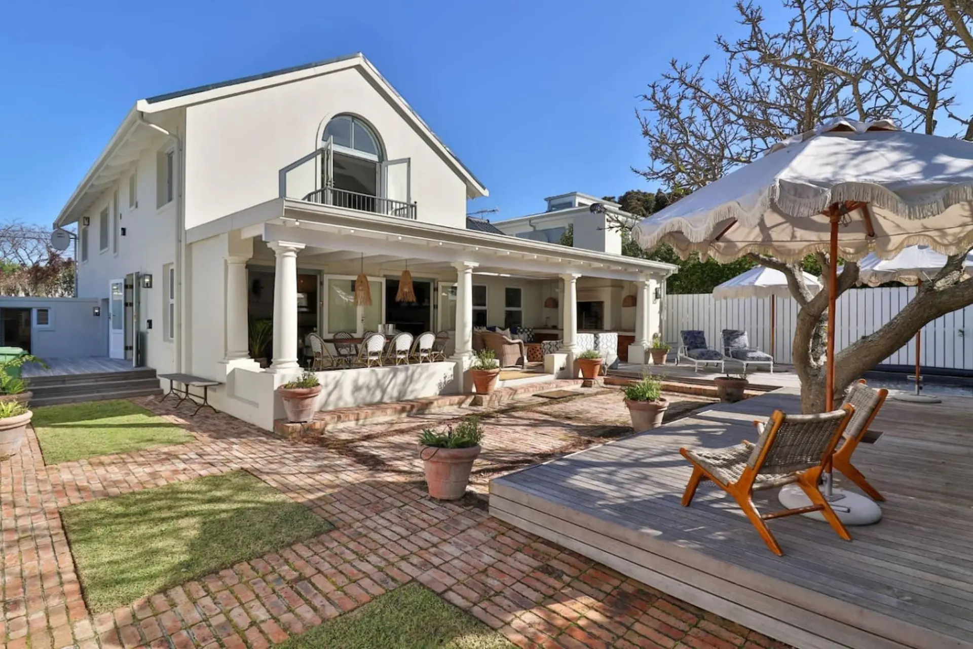A white house with a patio and deck, featuring outdoor seating, umbrellas, and lush greenery.
