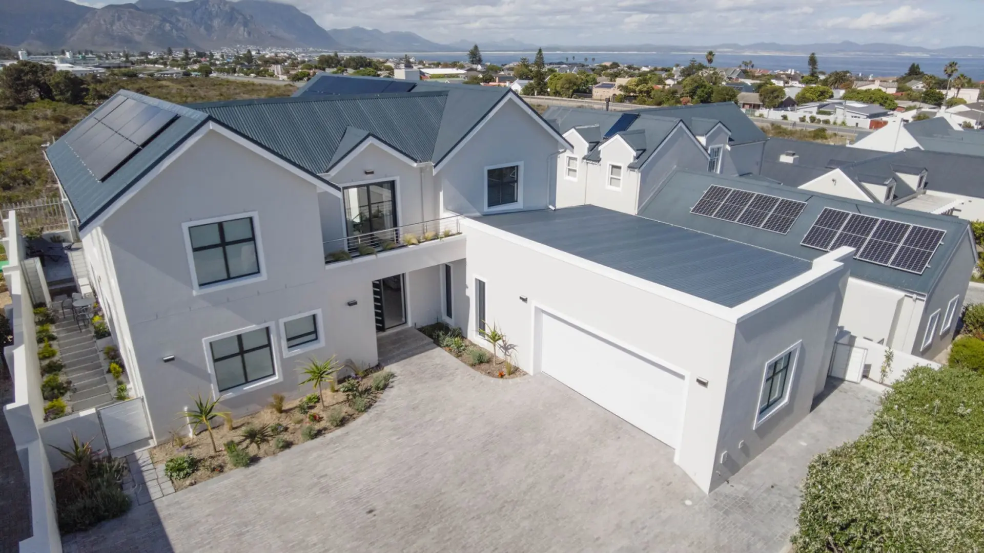 Modern house with solar panels on a blue-grey metal roof, a driveway, and a scenic coastal backgroun