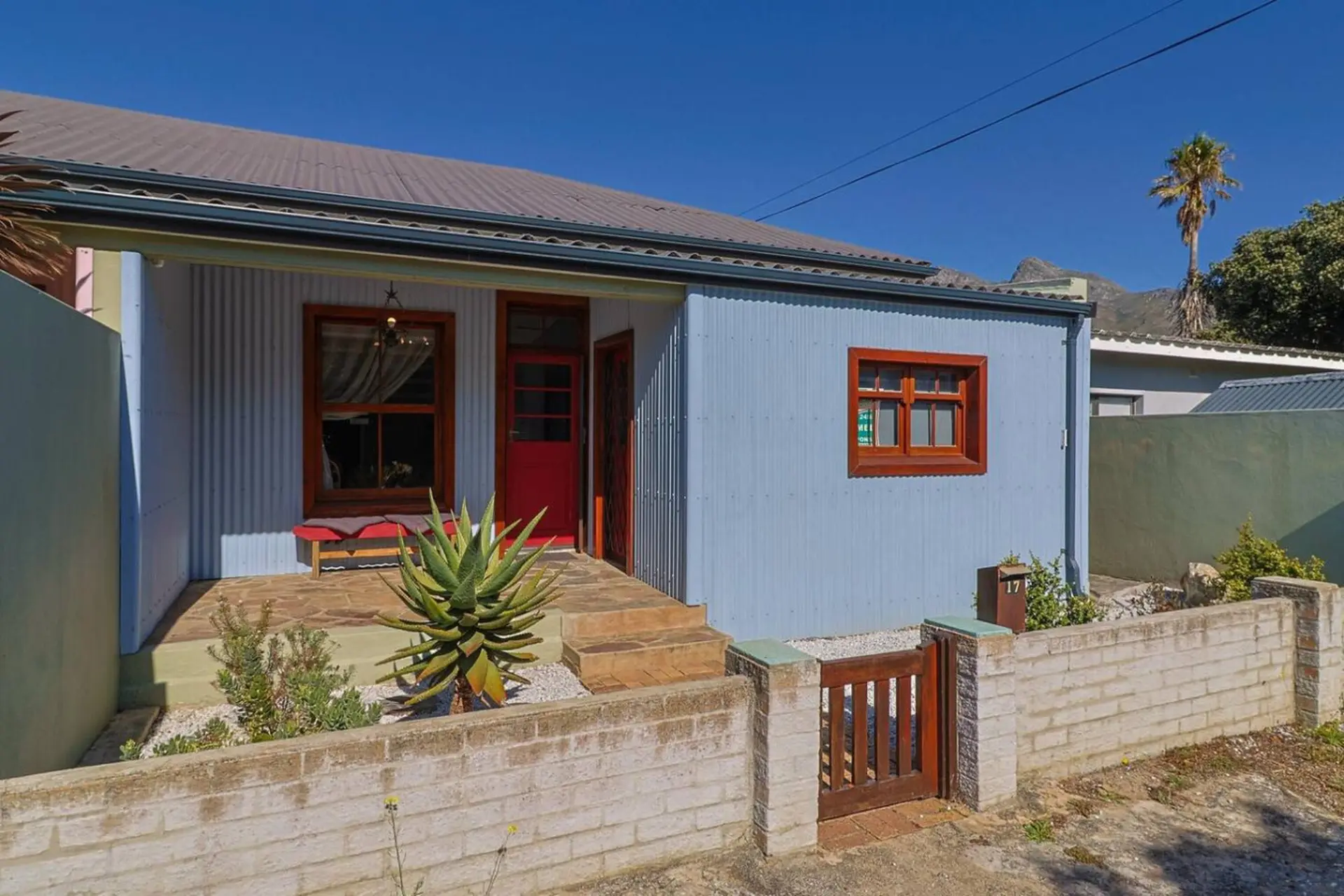 A light blue corrugated metal house with a red door and wooden window frames.