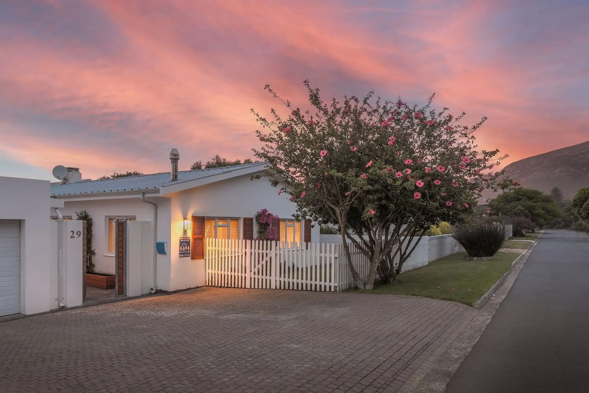 A white house with a garden fence and a blooming tree under a pink sky.