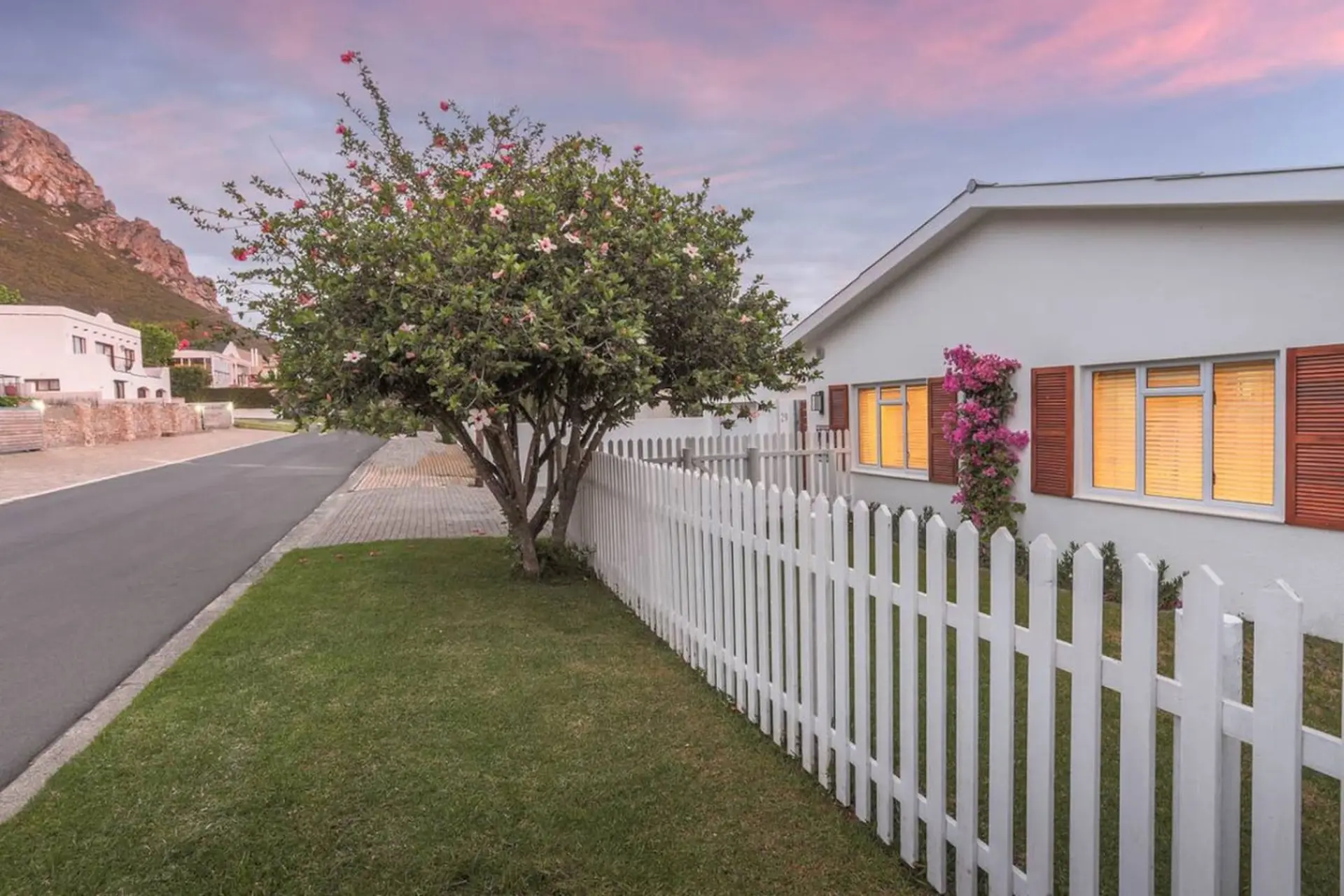 A white picket fence borders a grassy yard with a tree and a house.
