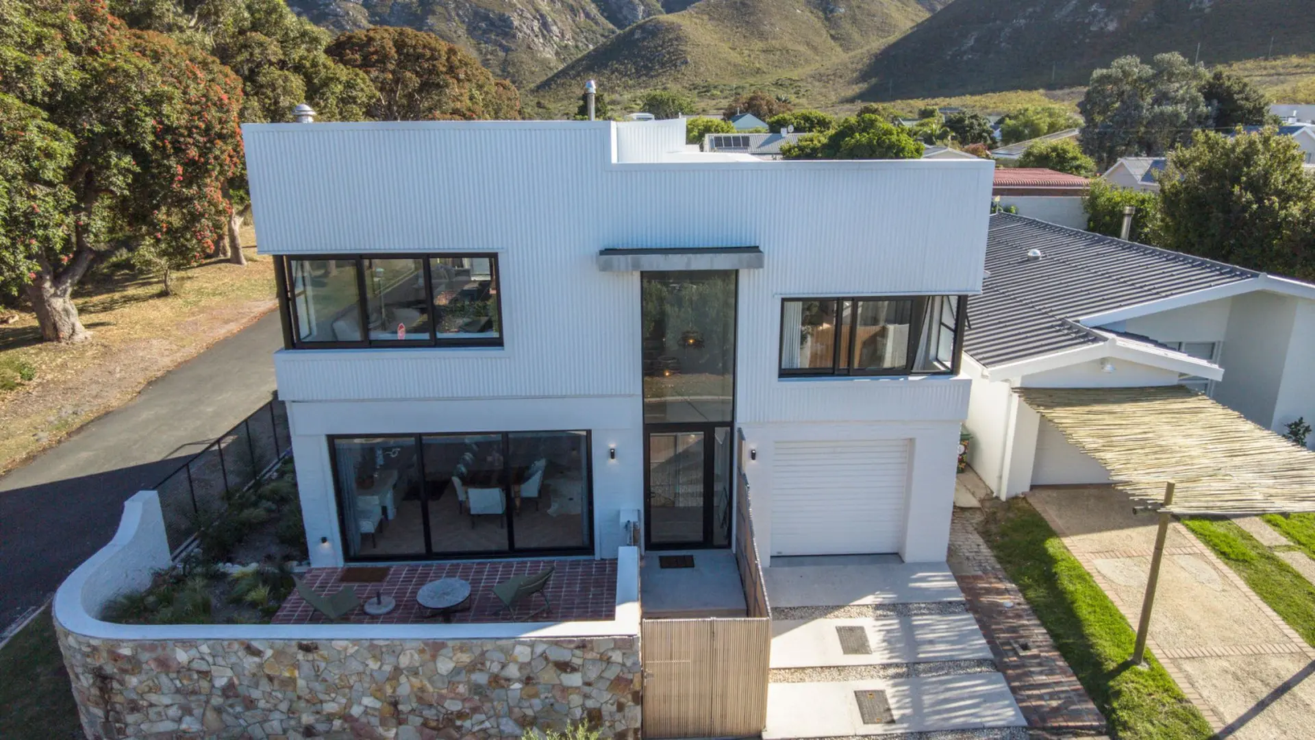 Modern white house with large windows and a corrugated metal exterior, set against a mountain backdr