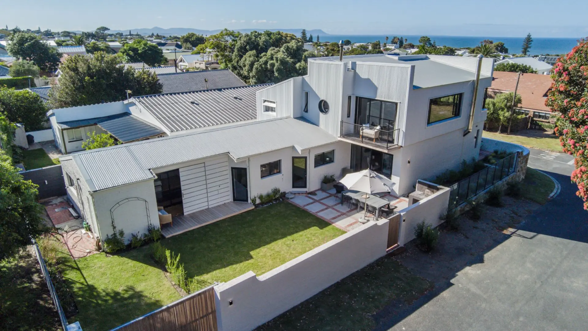 An aerial view of a modern, white house with a corrugated metal roof and a large backyard.