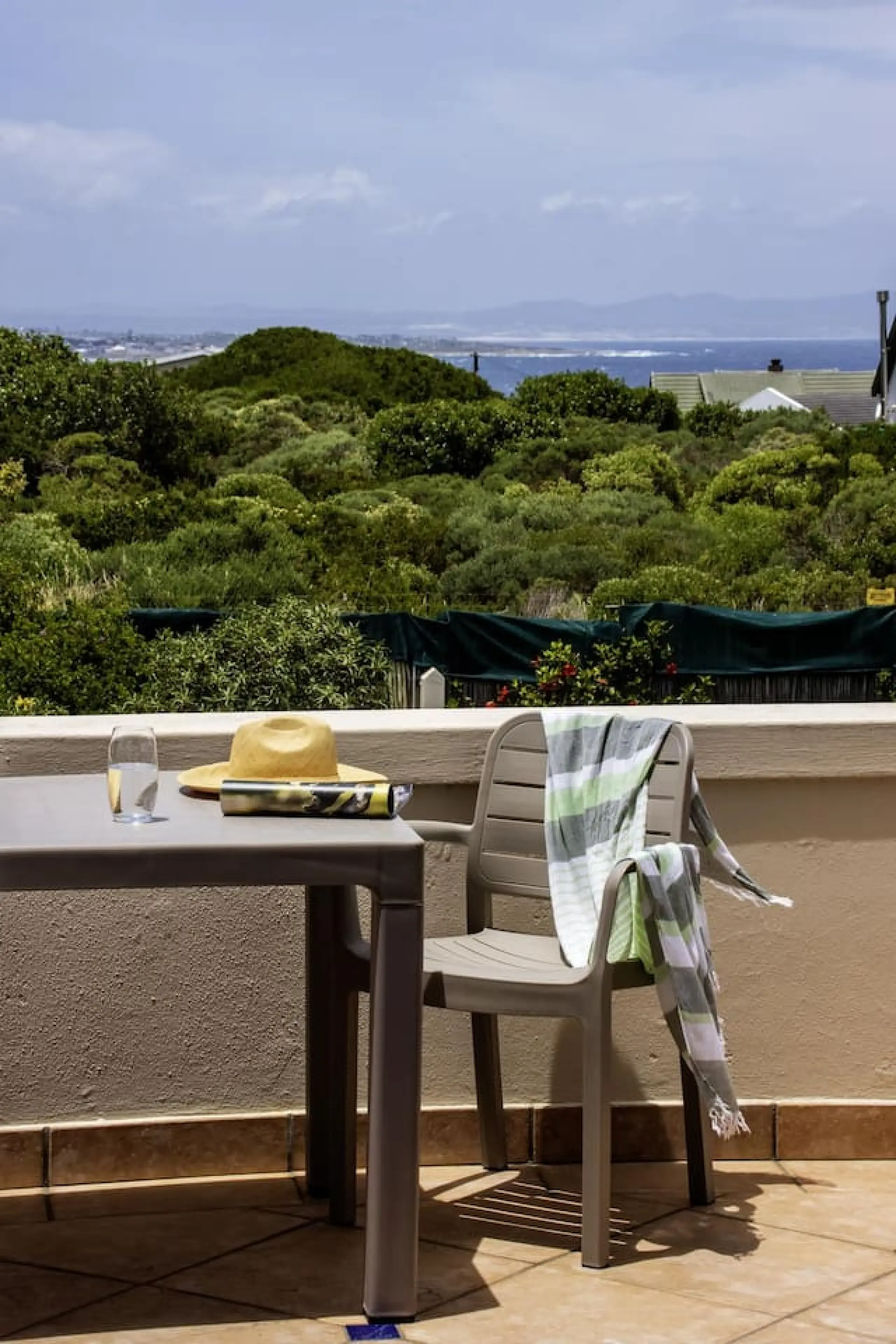 A table and chair with a hat and towel overlook a green landscape and the ocean.