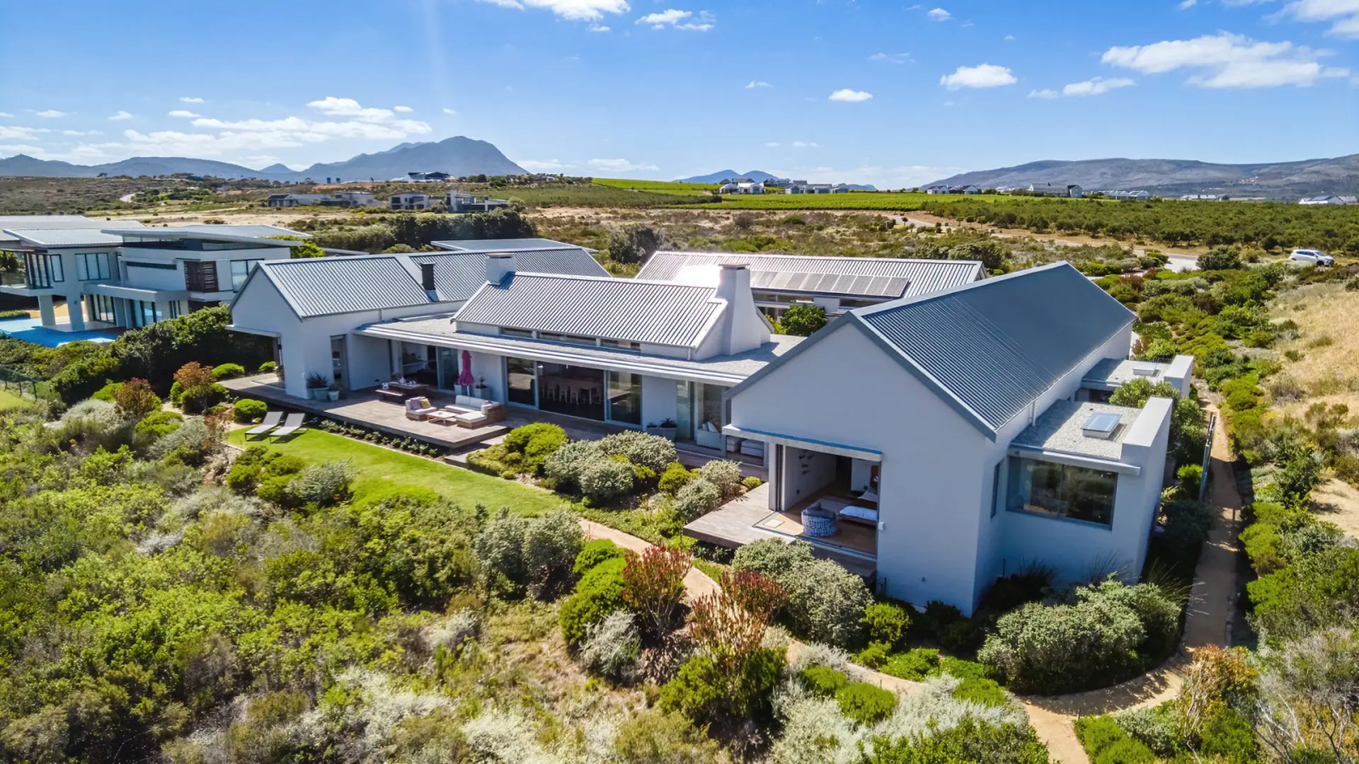 Modern white house with grey roofs and solar panels, surrounded by lush green landscaping.