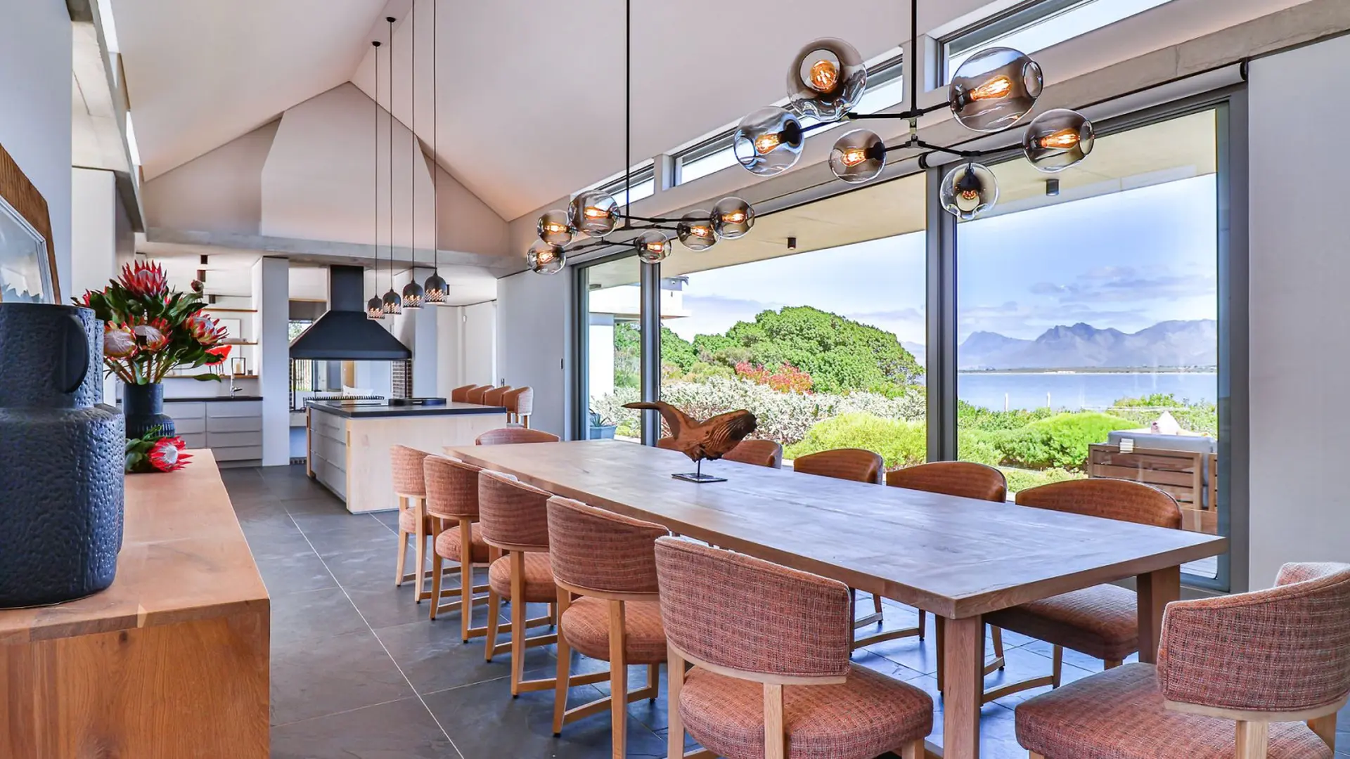 A modern dining room with a long wooden table and upholstered chairs, overlooking a scenic waterfron