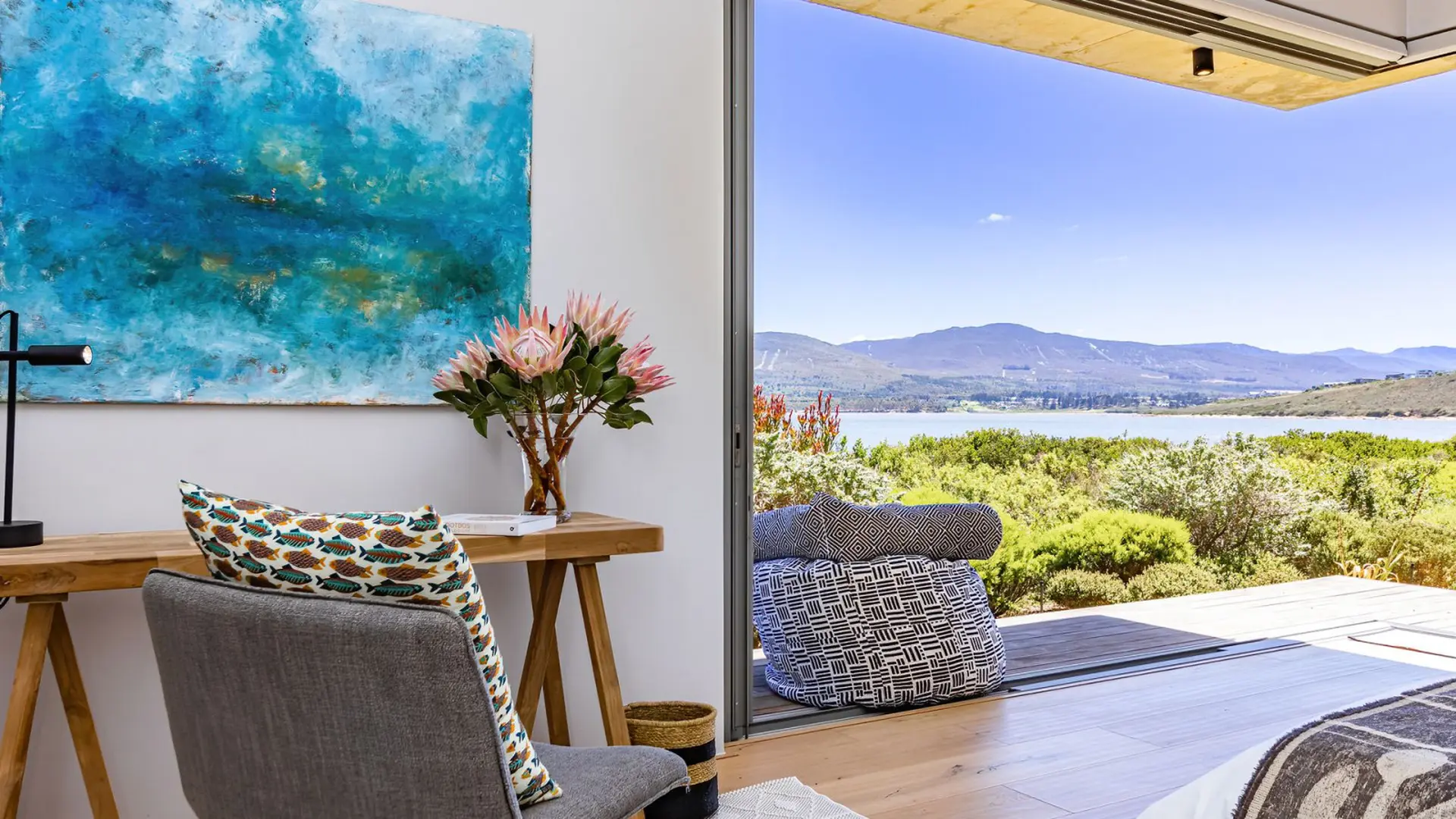 A desk with a painting, flowers, and a chair faces a view of a lake and mountains.