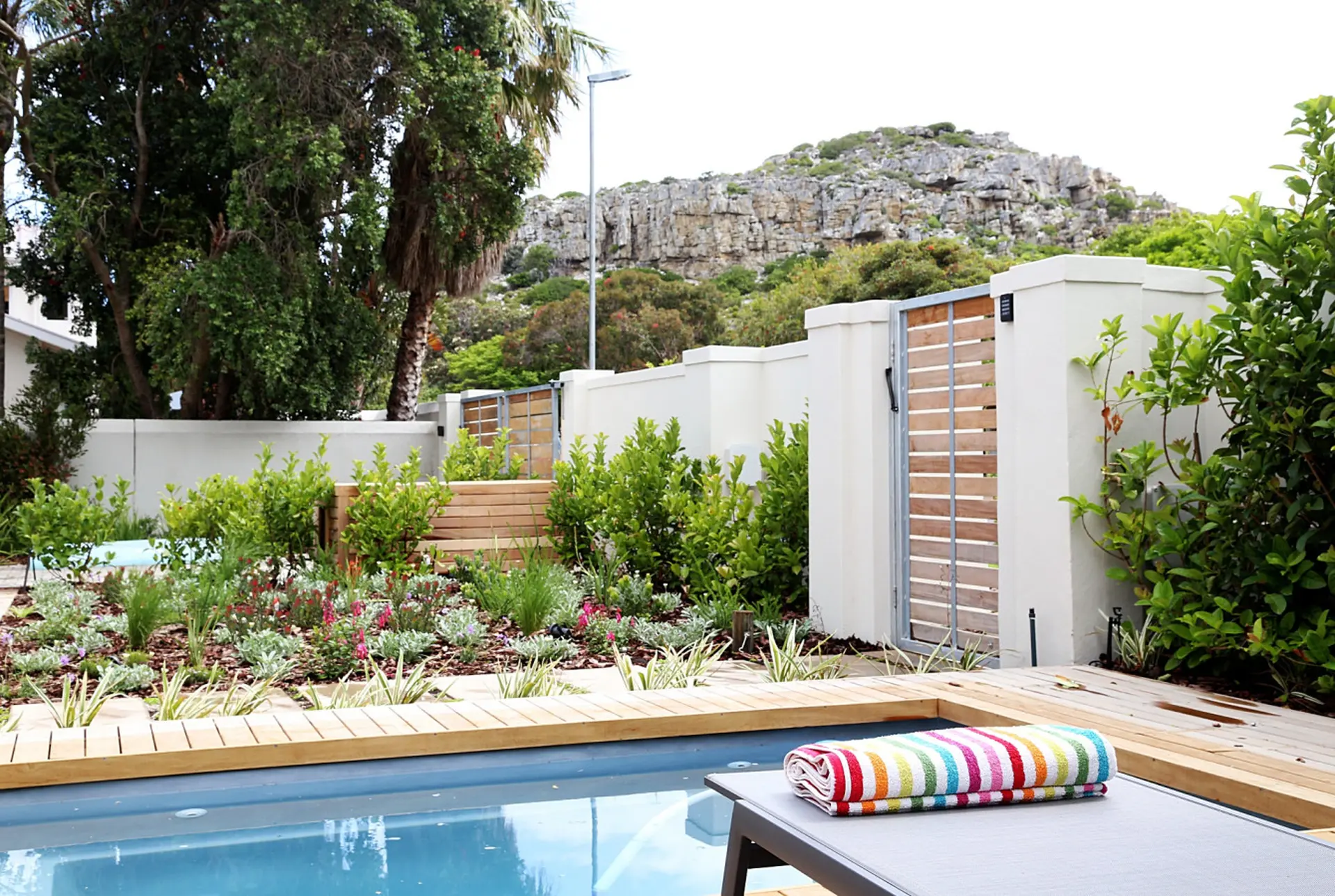 A poolside lounge chair with a striped towel sits by the pool, with a garden and rock cliff in the b