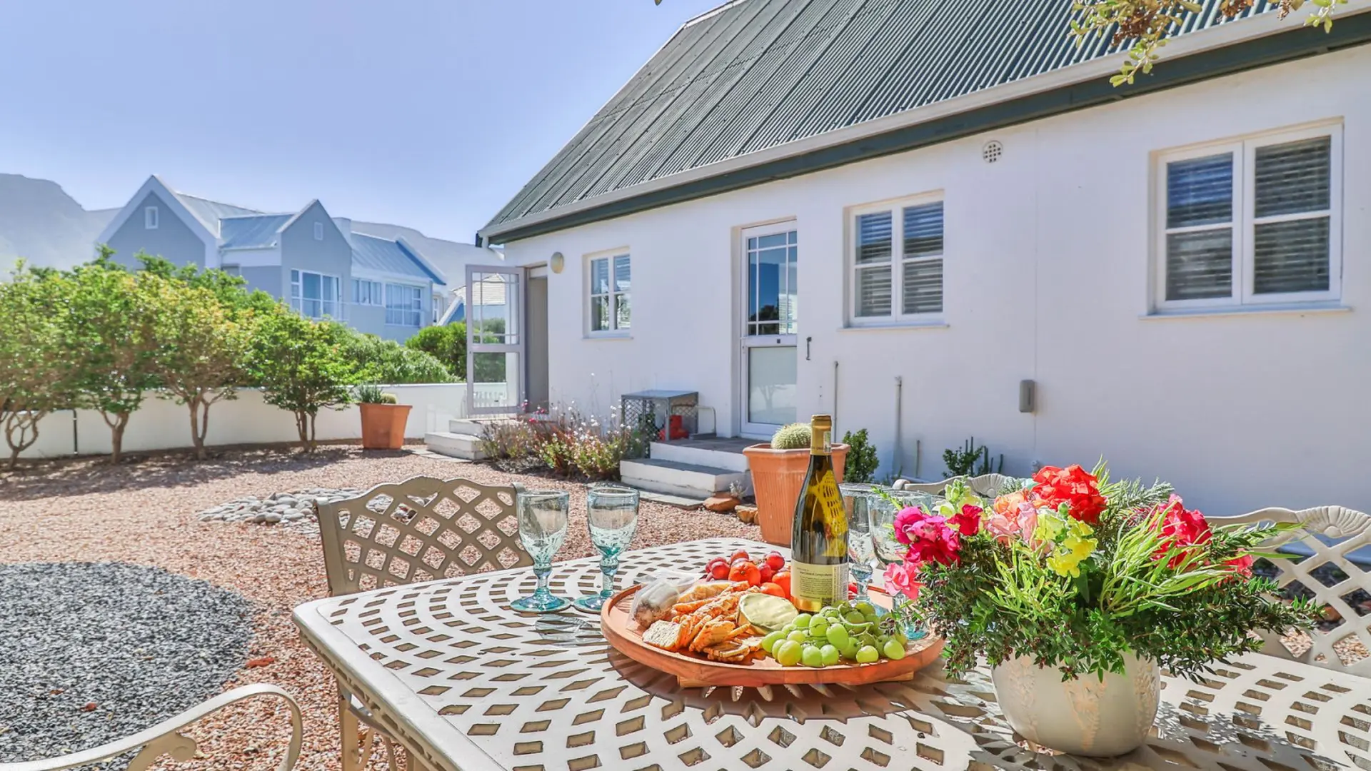 A table laden with food, wine, and flowers sits in a sunny courtyard.