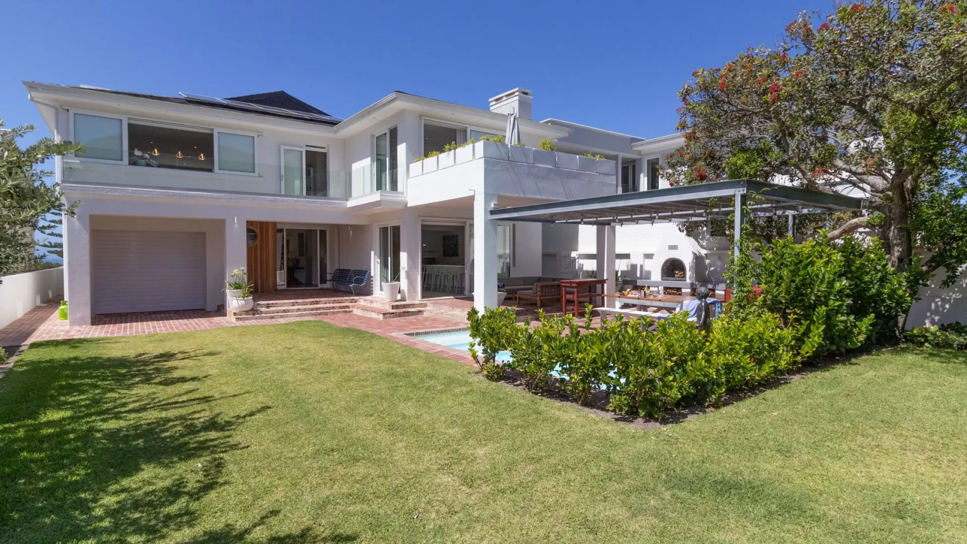 A modern, white, multi-story house with a garage, patio with seating, and a swimming pool in the gar
