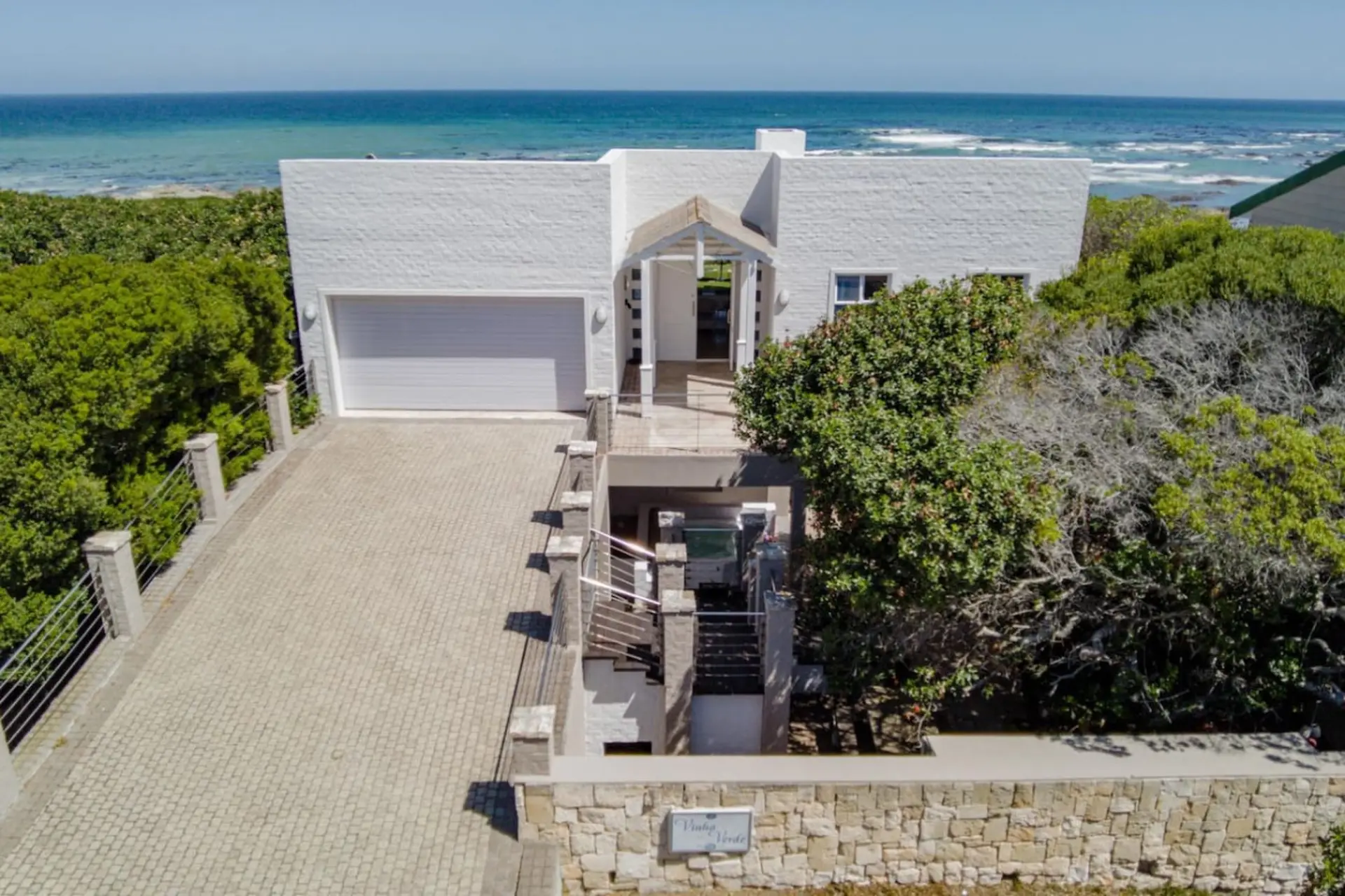 A white house with a garage and a paved driveway sits on a bluff overlooking the ocean.