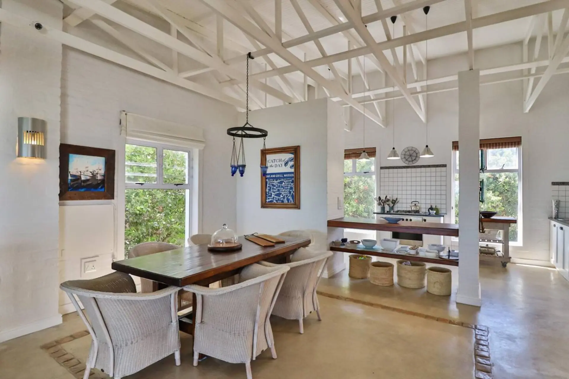 A sunlit dining room with a wooden table and wicker chairs, leading to a kitchen with white tiled wa