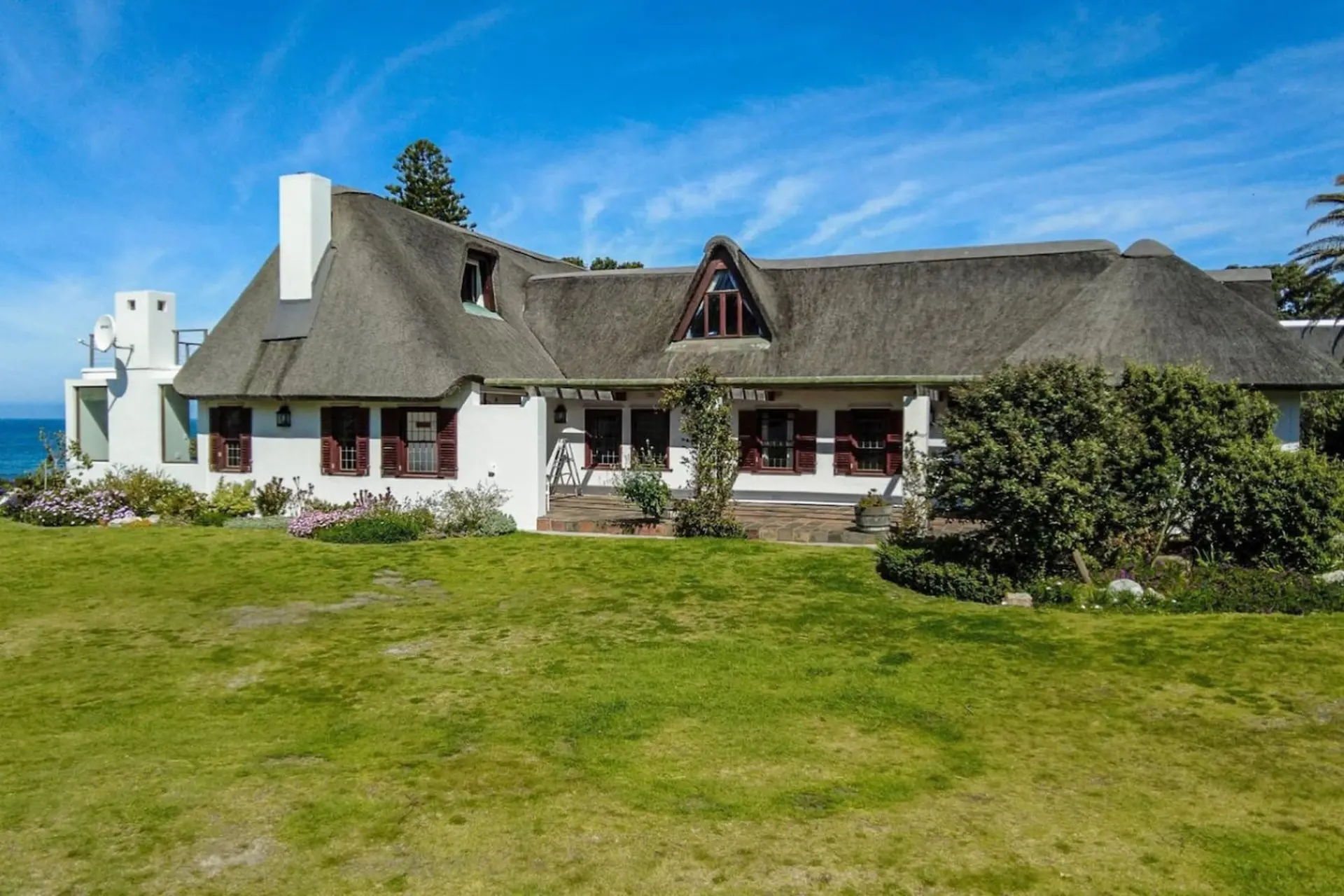 A white house with a thatched roof and brown shutters sits on a grassy lawn under a blue sky.