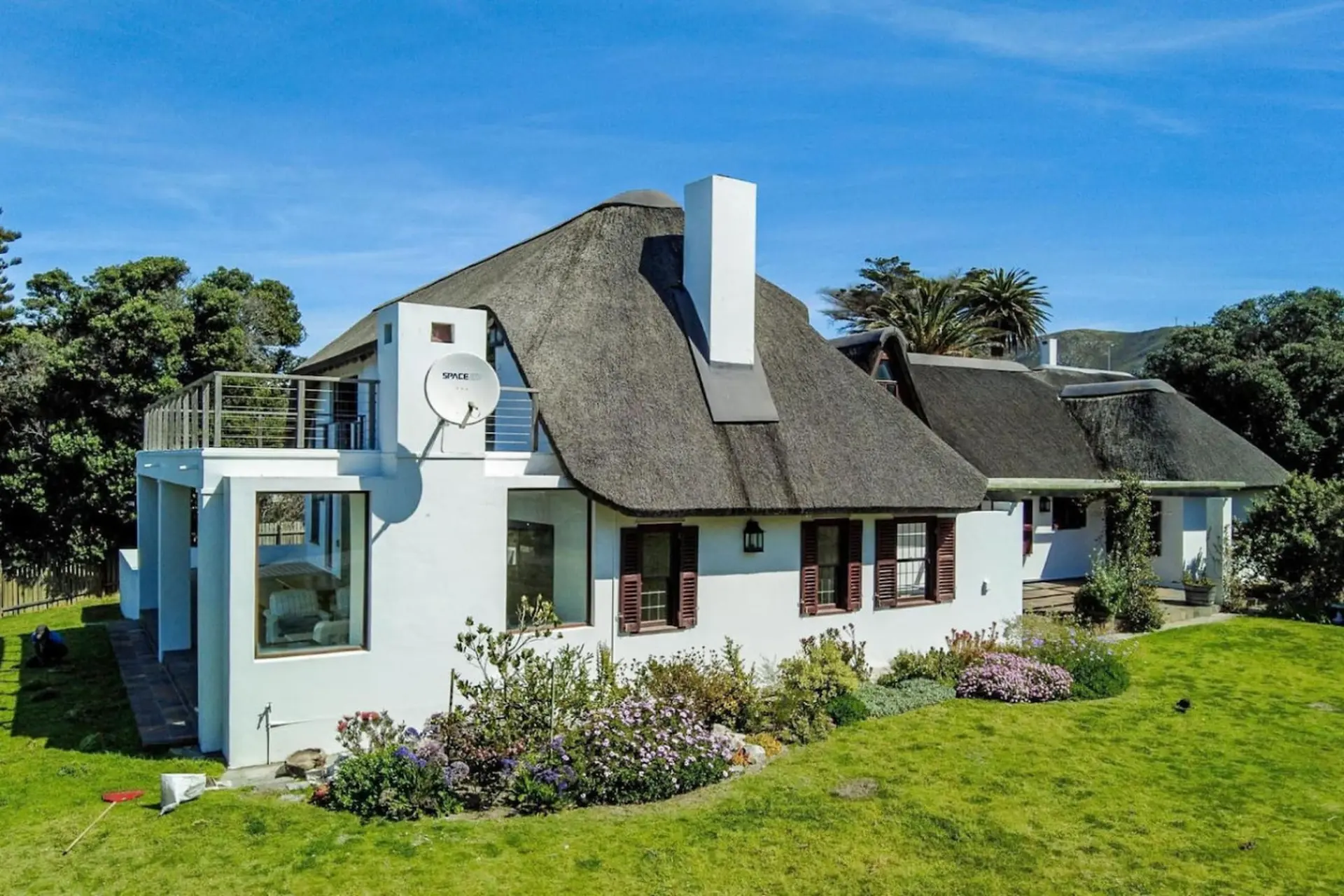 A white house with a thatched roof, balcony, and garden under a blue sky.
