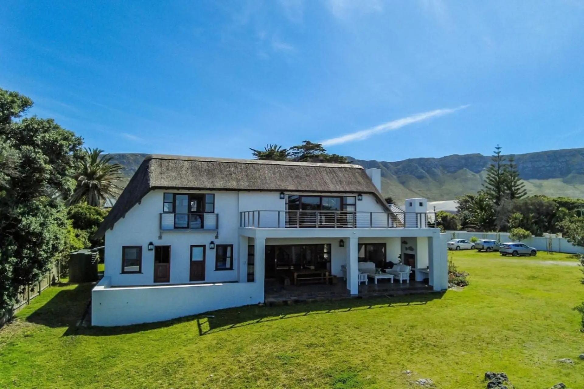 A white house with a thatched roof sits on a grassy lawn with mountains in the background.