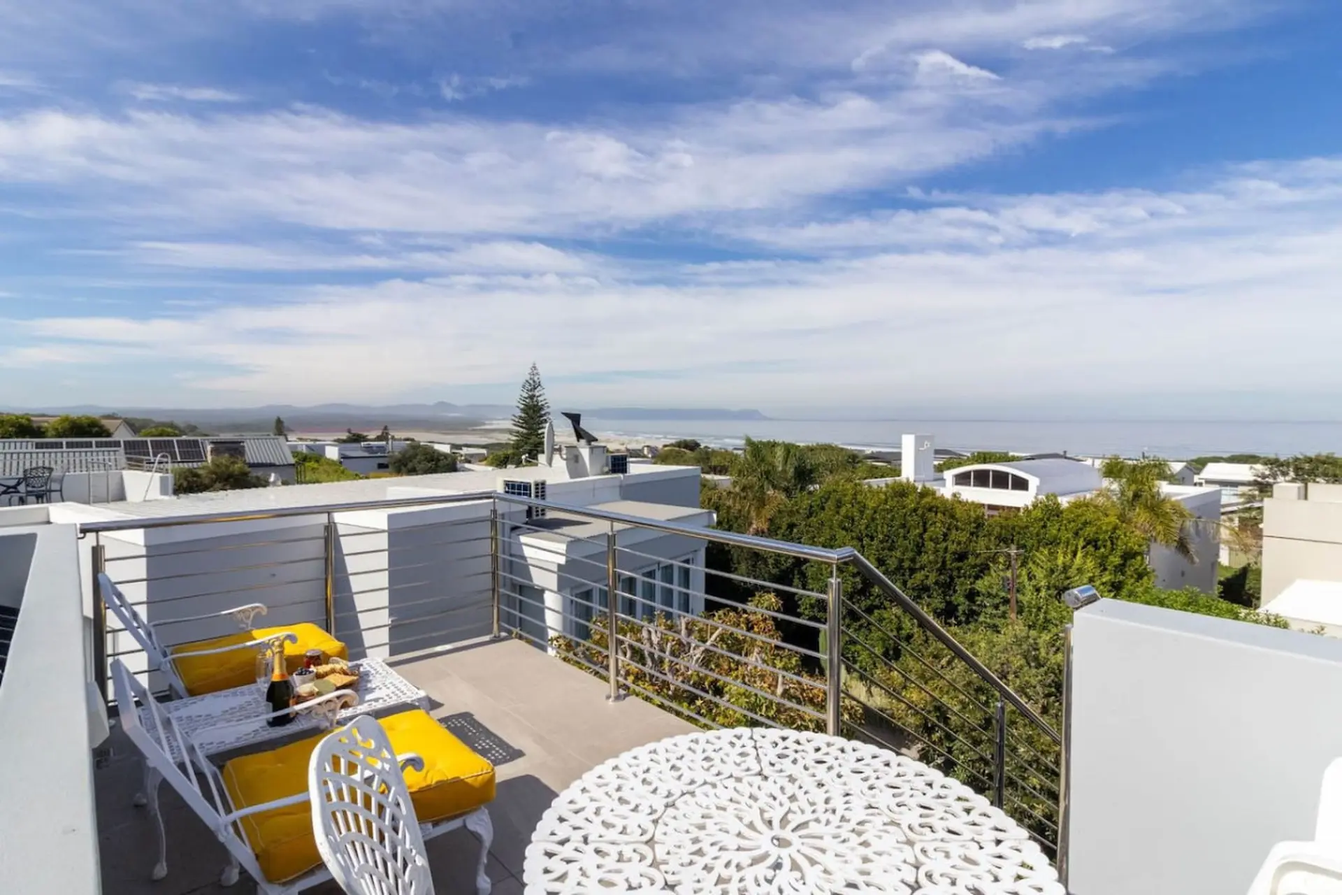 A white table and chairs with yellow cushions on a balcony overlook the ocean.
