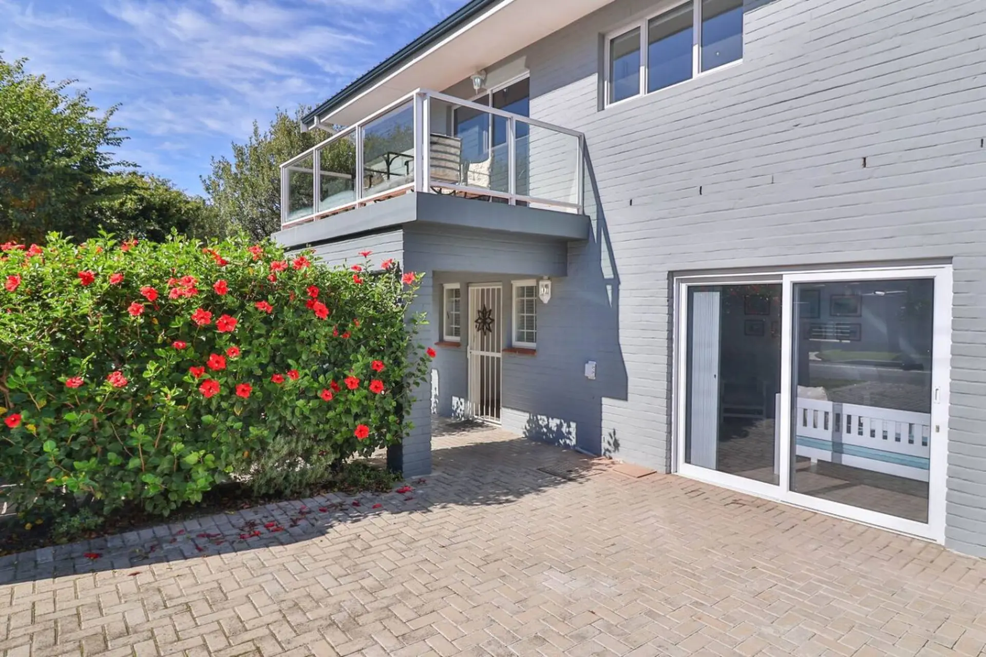 Modern grey brick house with balcony, sliding glass doors, and vibrant red hibiscus flowers.