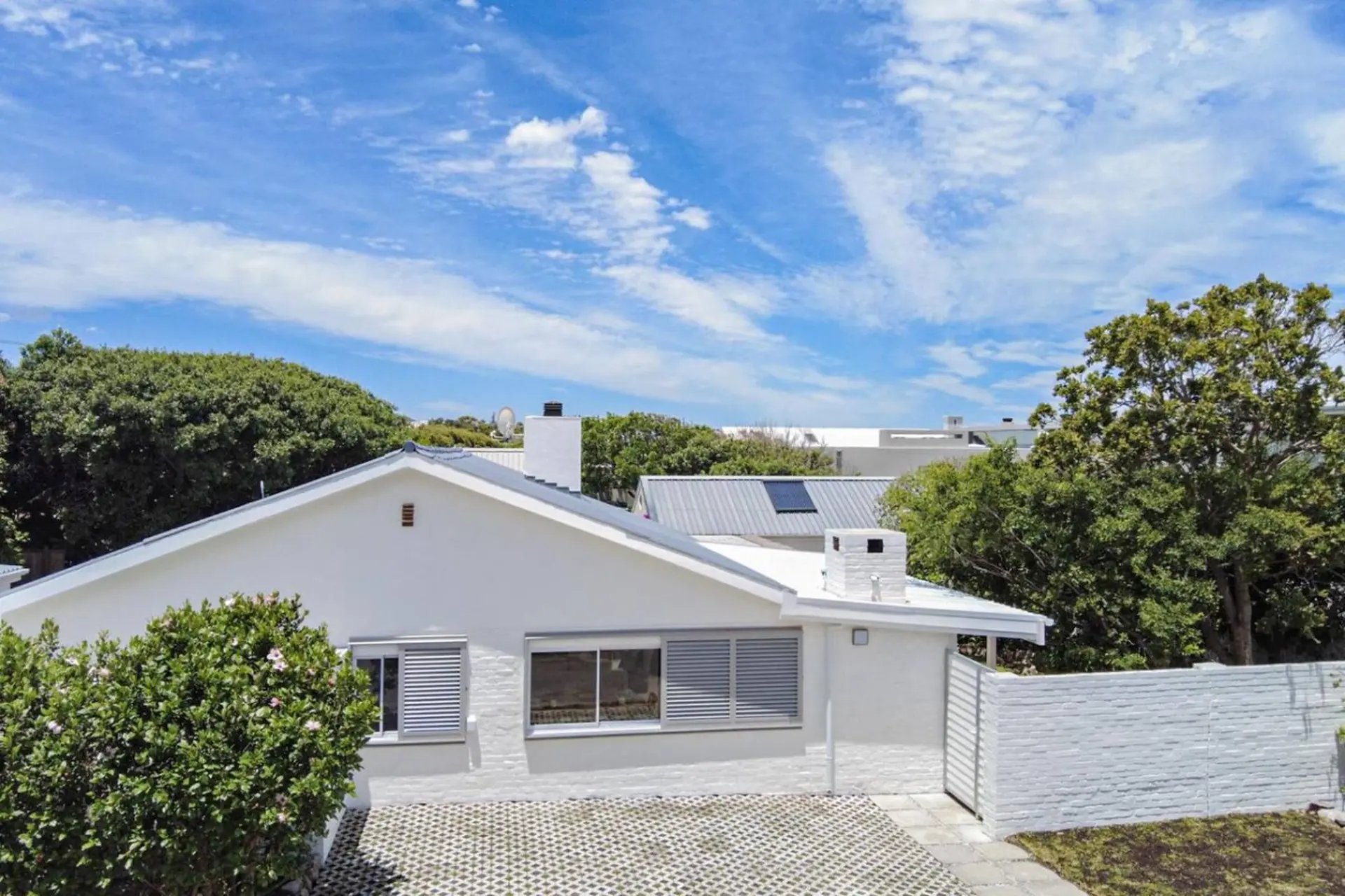 A white house with grey shutters and a white brick wall under a bright blue sky.