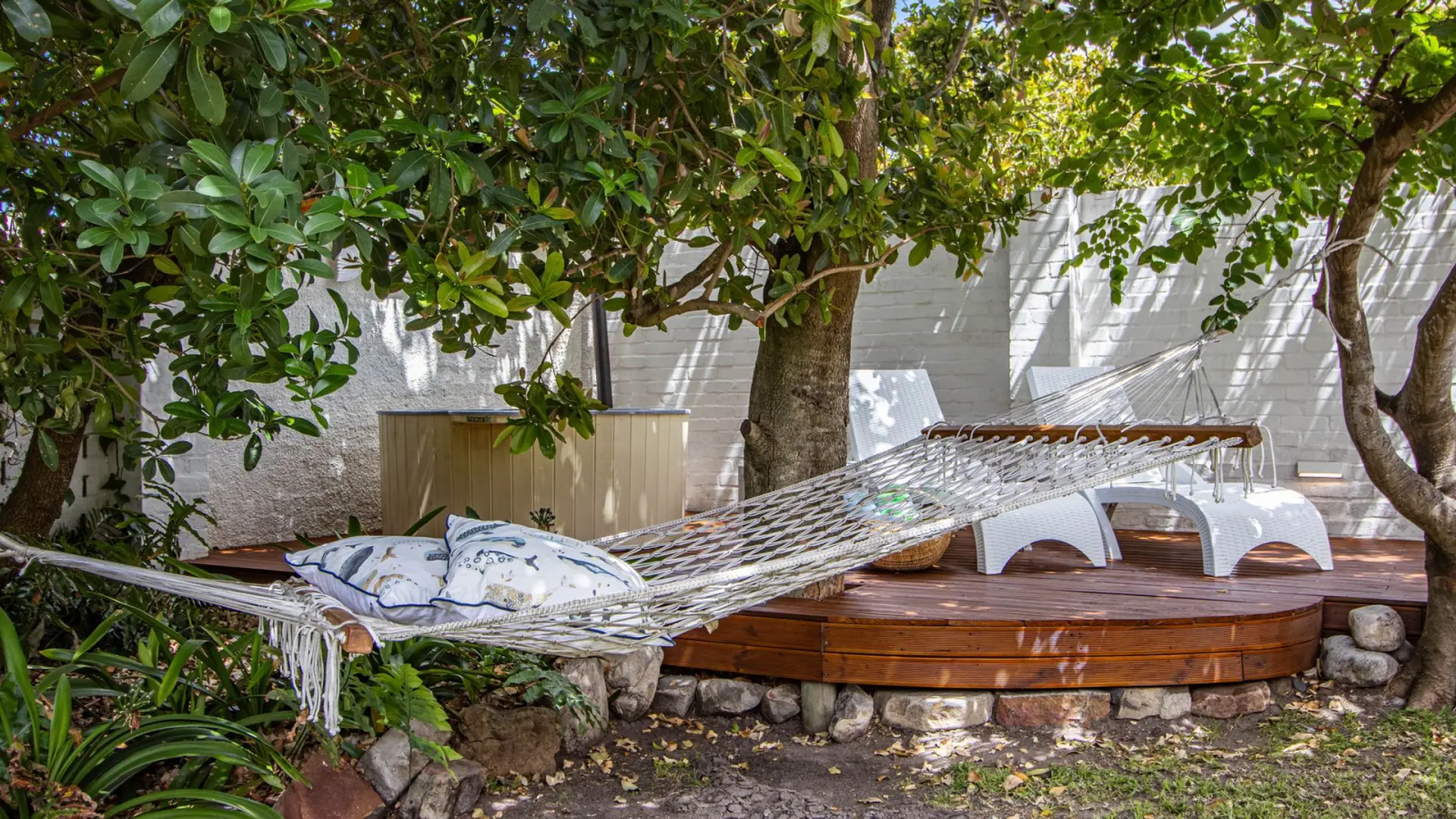 Hammock hanging in a backyard with trees, white brick wall, and deck.