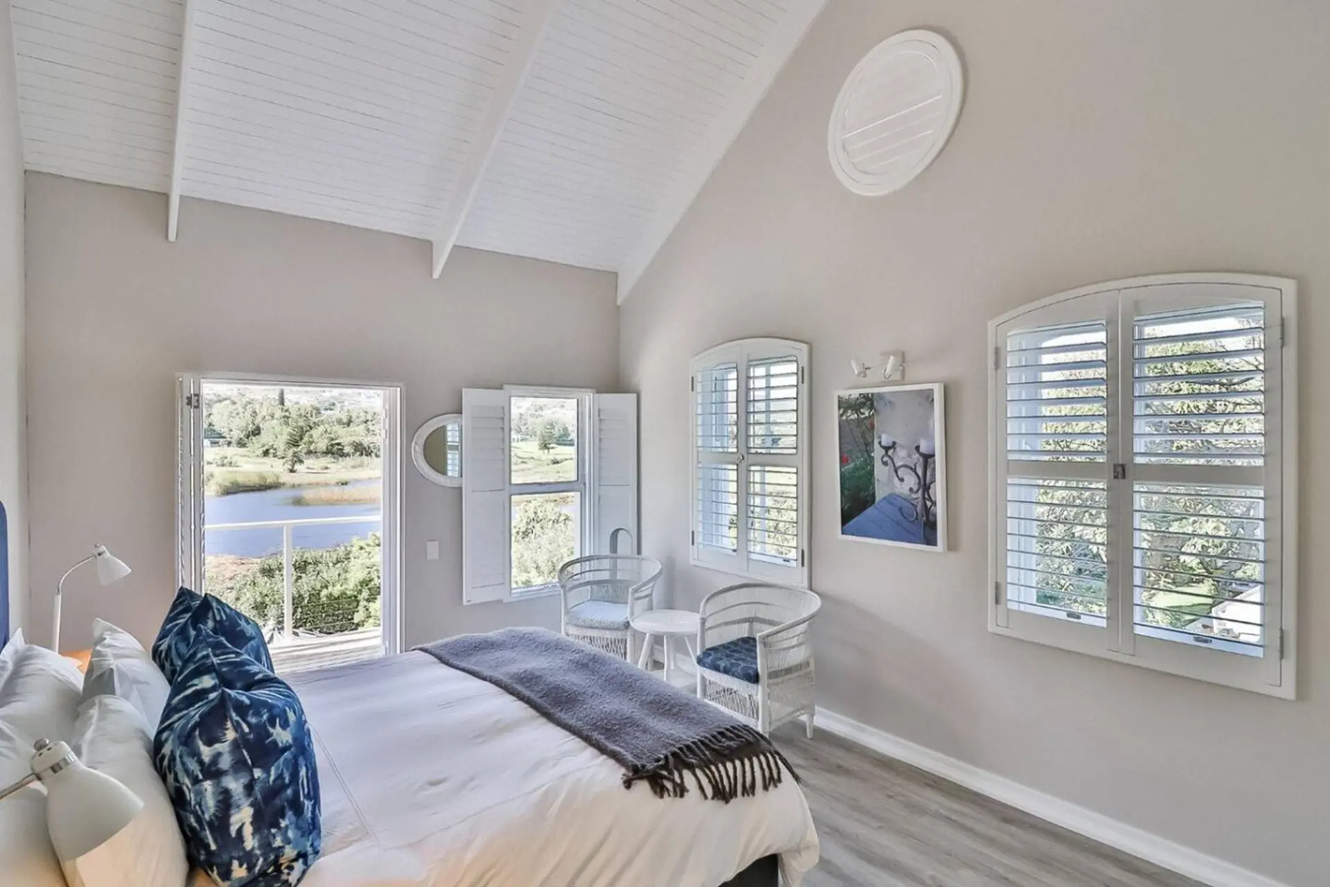 A bedroom with white shutters, a king-size bed, and a view of a lake.