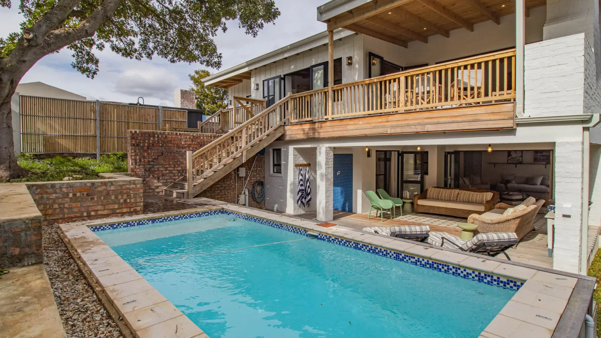 A bright blue swimming pool is in the foreground with a modern house and wooden deck in the backgrou