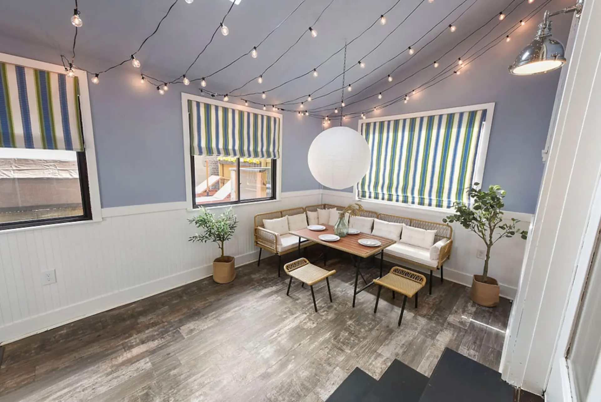 A light blue room with string lights, striped window coverings, and a dining set with rattan furnitu