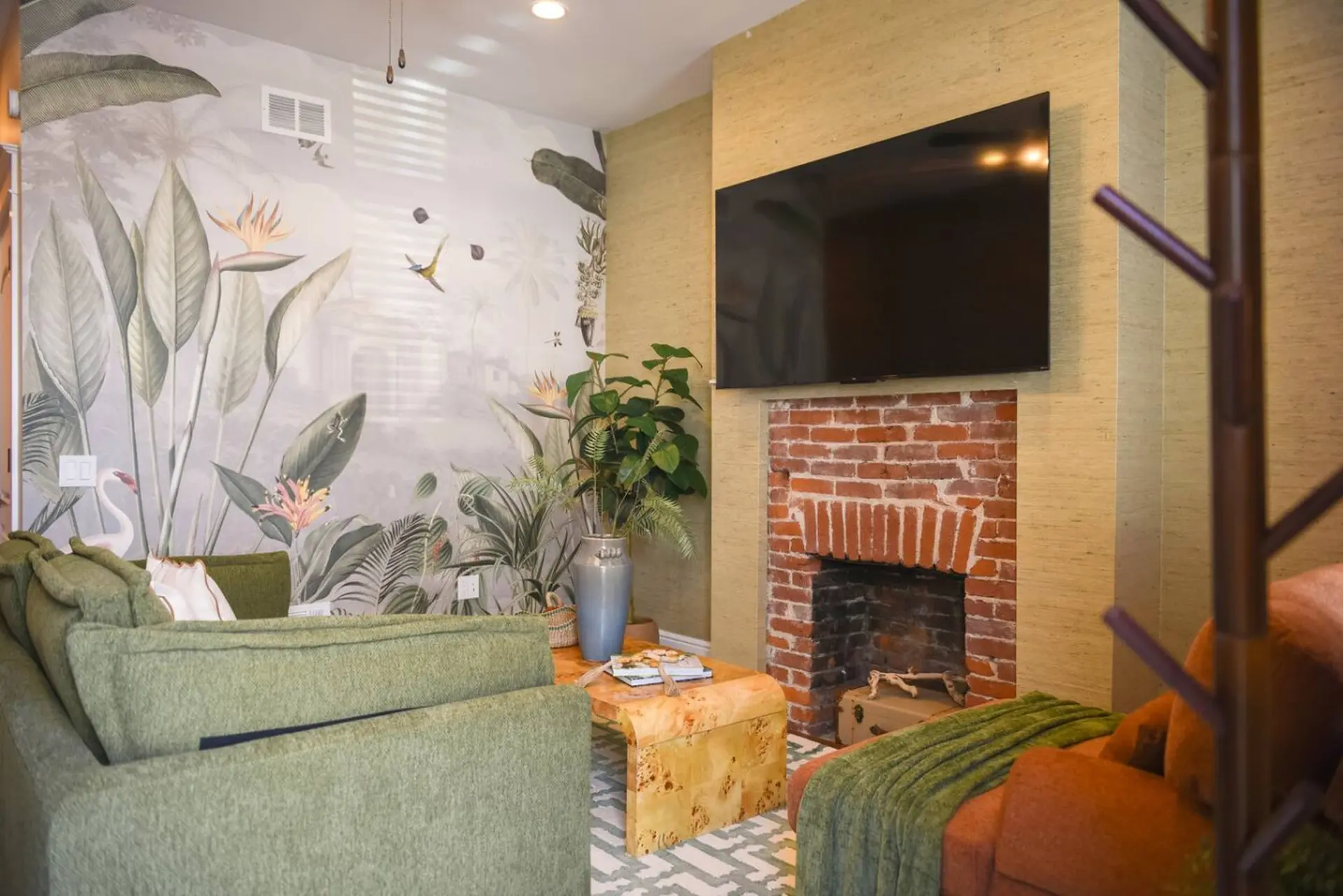 Living room with tropical wallpaper, green sofas, and a brick fireplace with a mounted TV.