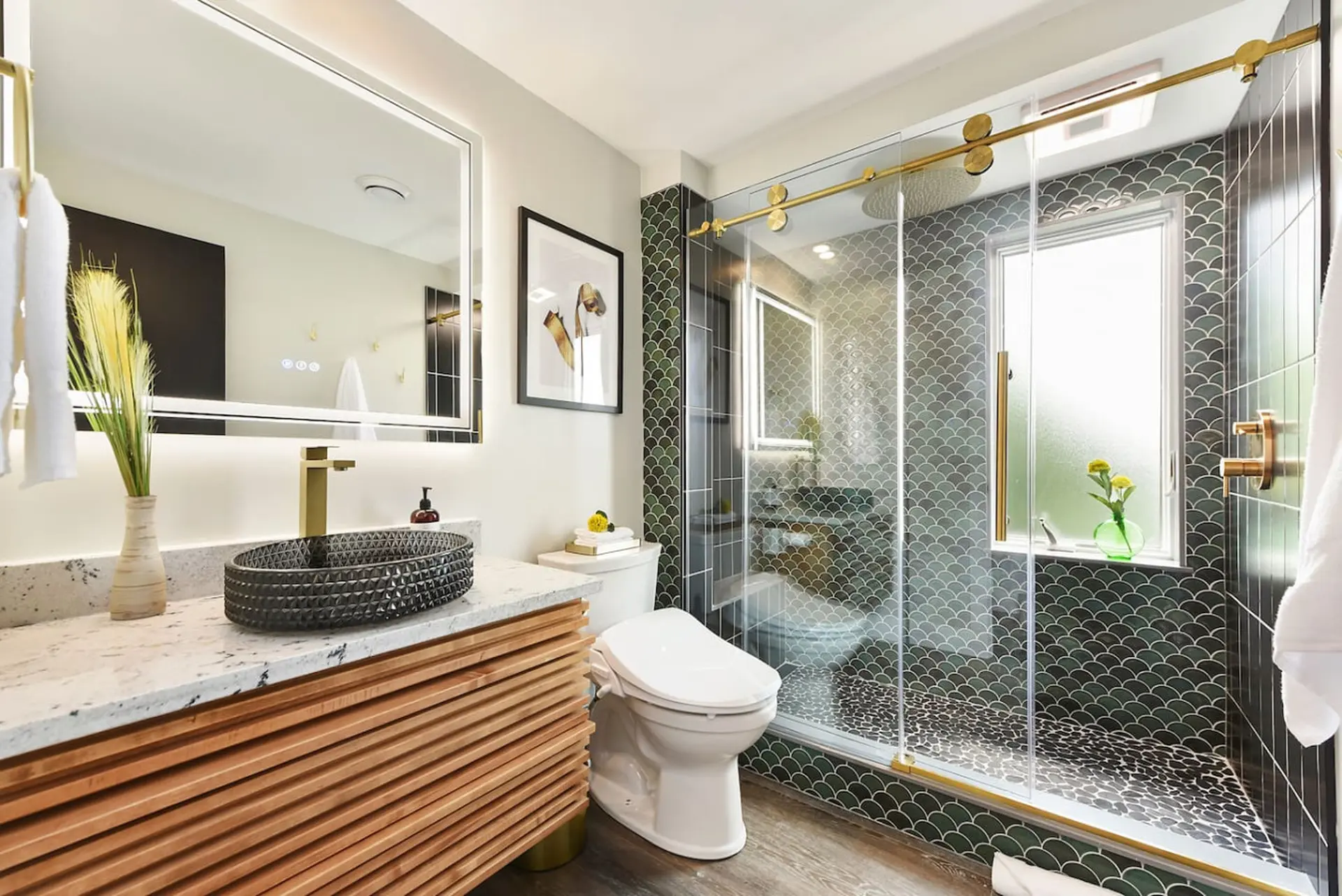 Modern bathroom with a wooden vanity, gray scale tile shower, and gold fixtures.