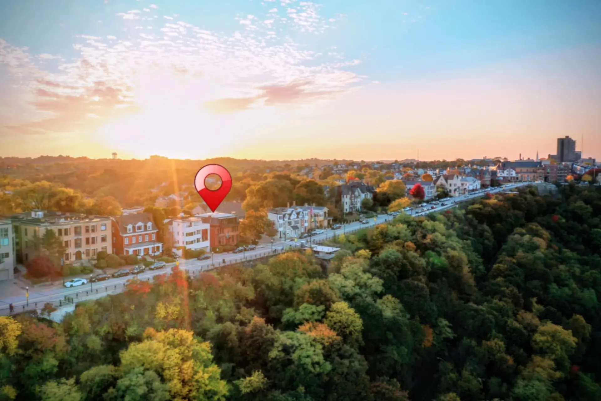 Drone view of a city skyline with fall foliage and a red location pin during sunset.