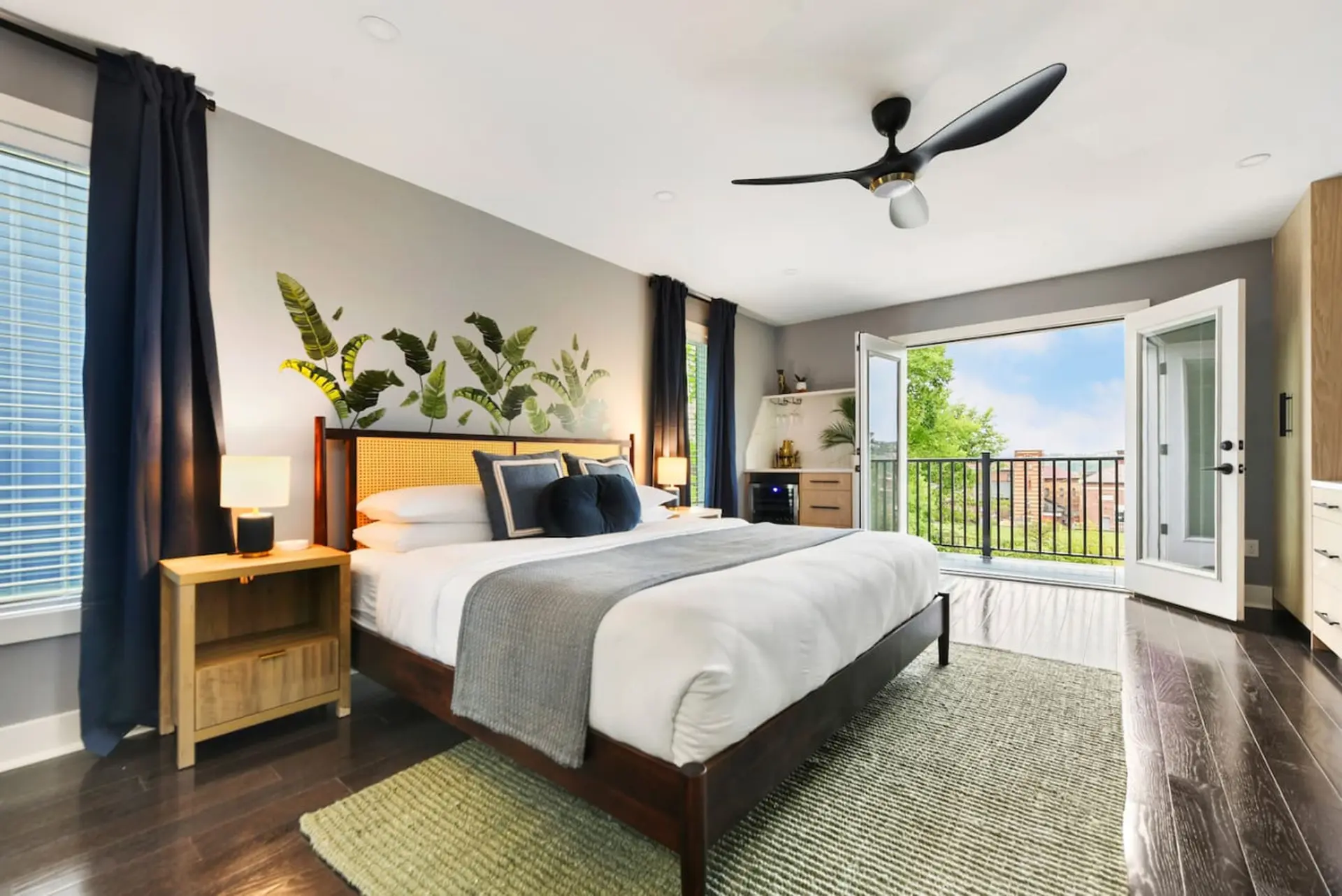 A king-size bed with white linens and a gray throw is in a bedroom with a tropical leaf mural and ba