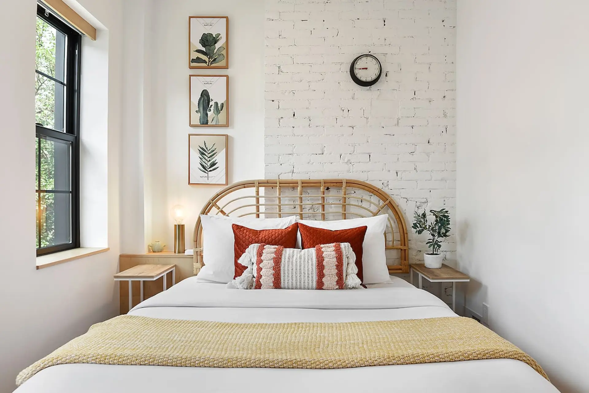 A bedroom with a white brick wall, a rattan headboard, and botanical art.
