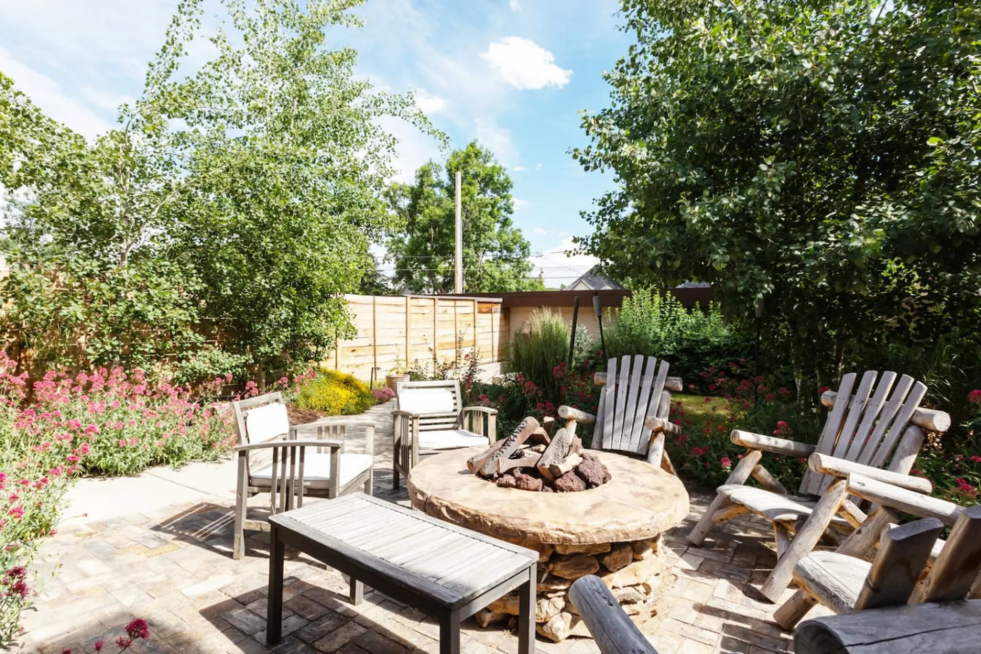 Outdoor patio with a stone fire pit, wooden chairs, and lush greenery.
