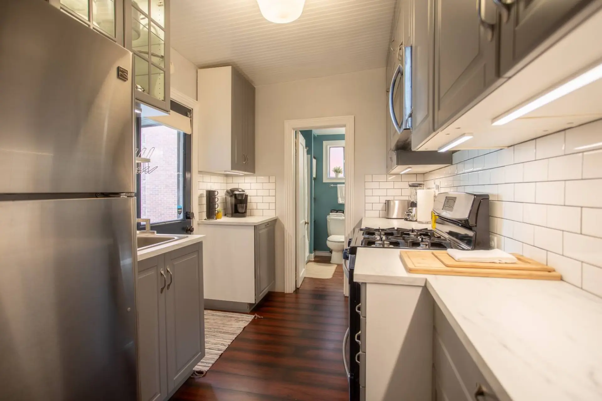 A narrow kitchen with white subway tiles, grey cabinets, and a stainless steel refrigerator.