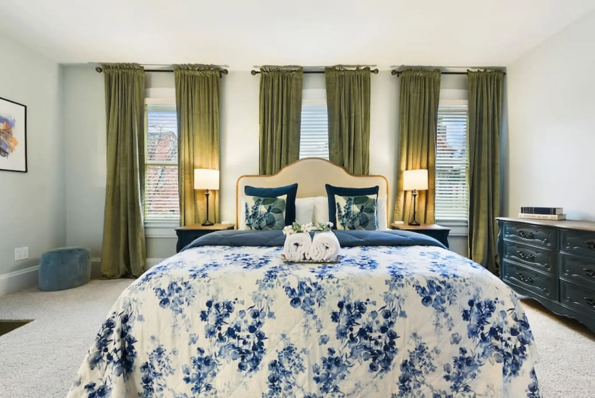 A bedroom with a king-sized bed and blue floral bedding, two nightstands with lamps, and green curta