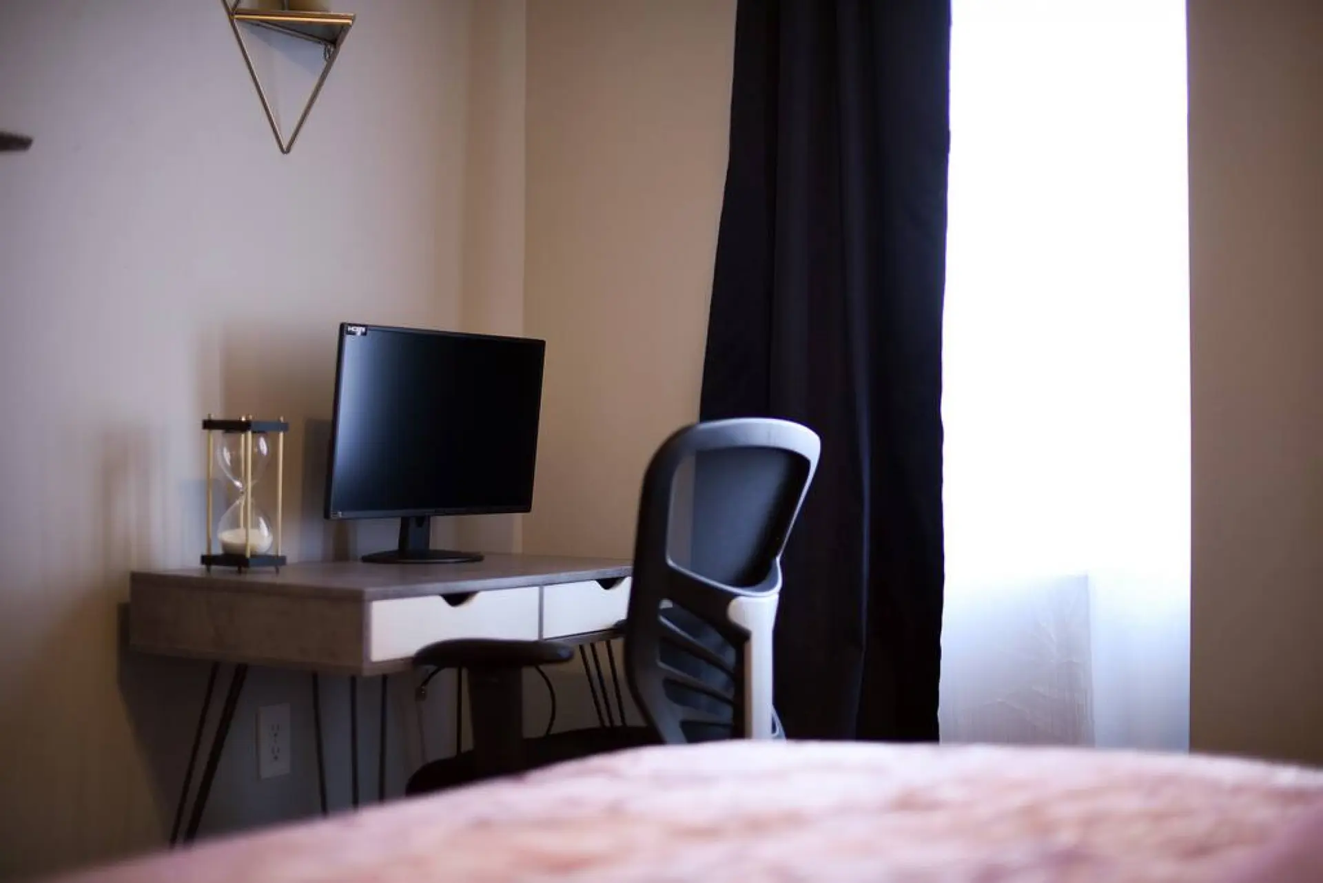 A desk with a computer monitor and hourglass sits in front of a black curtain and bright window.