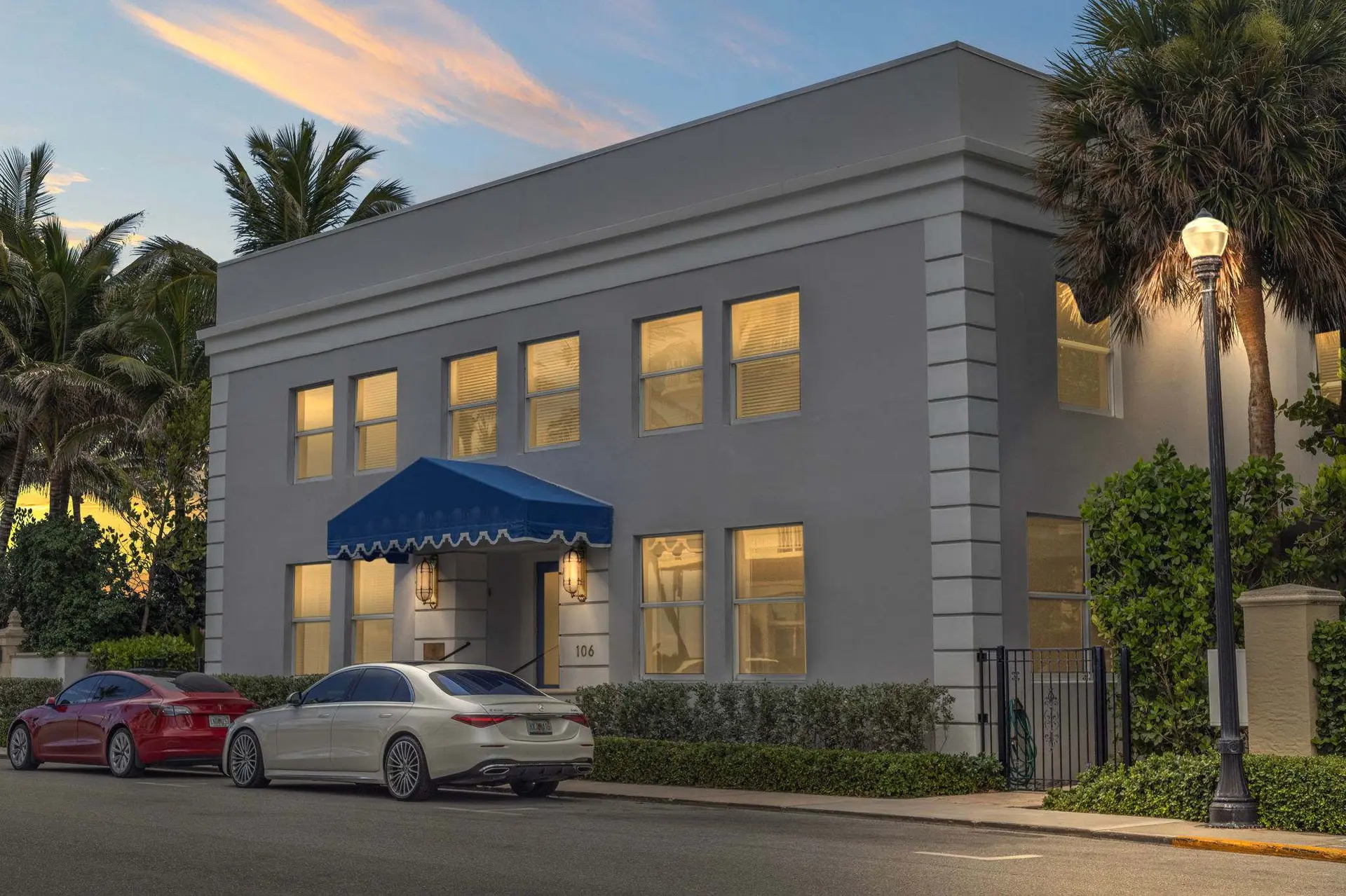 Luxury cars parked by a modern building with palm trees against a sunset sky.