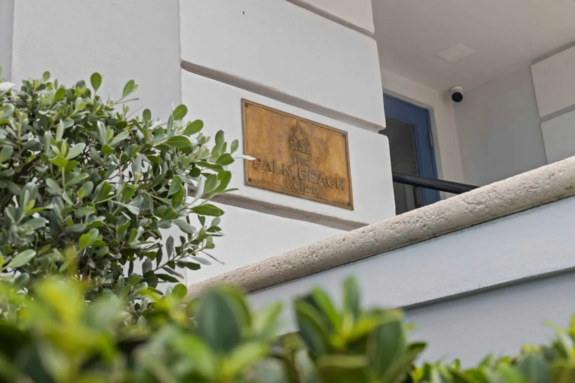A brass plaque on a white wall reads "The Palm Beach House."