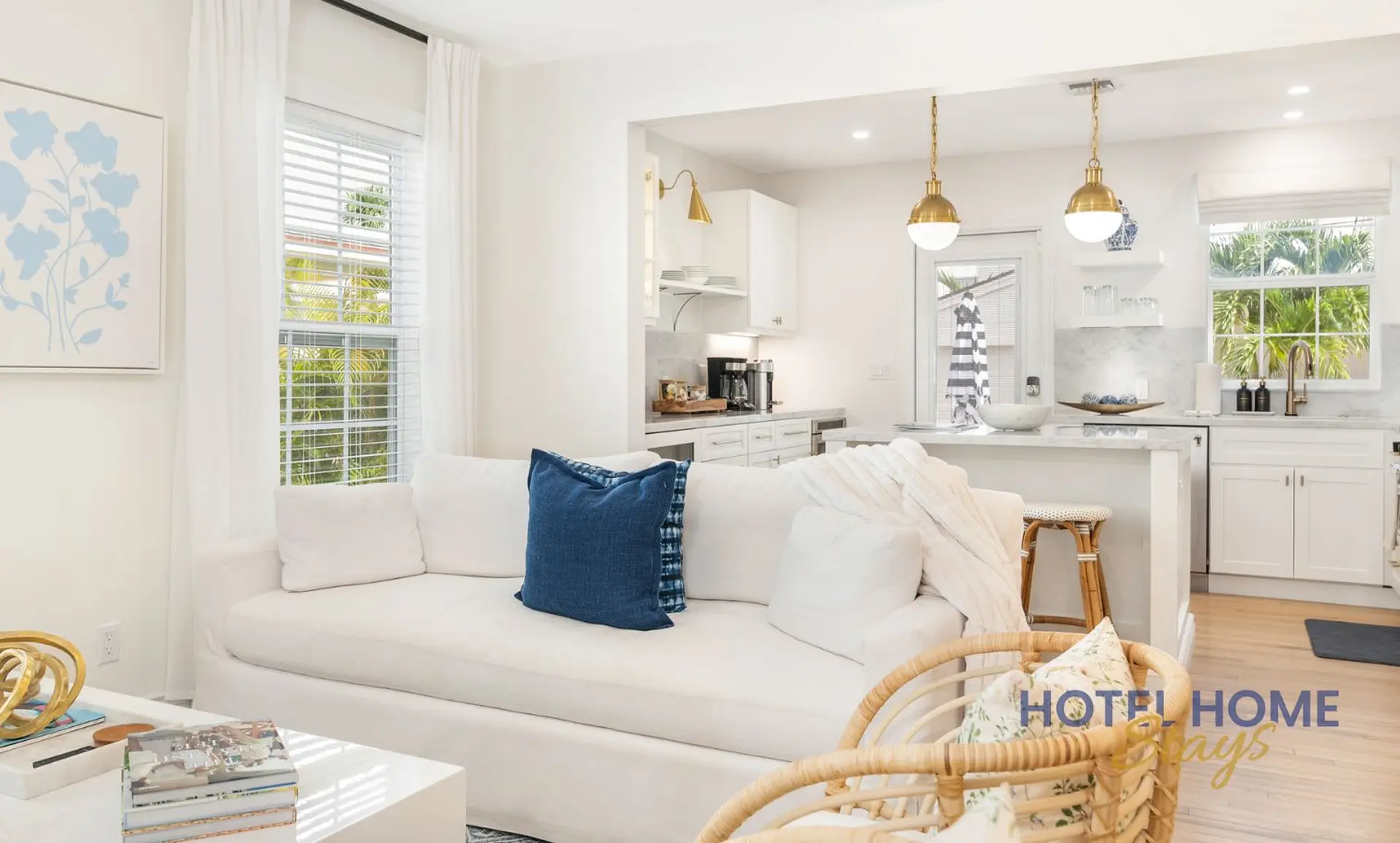 A white sofa with blue pillows sits in a bright living room, adjacent to a modern kitchen.