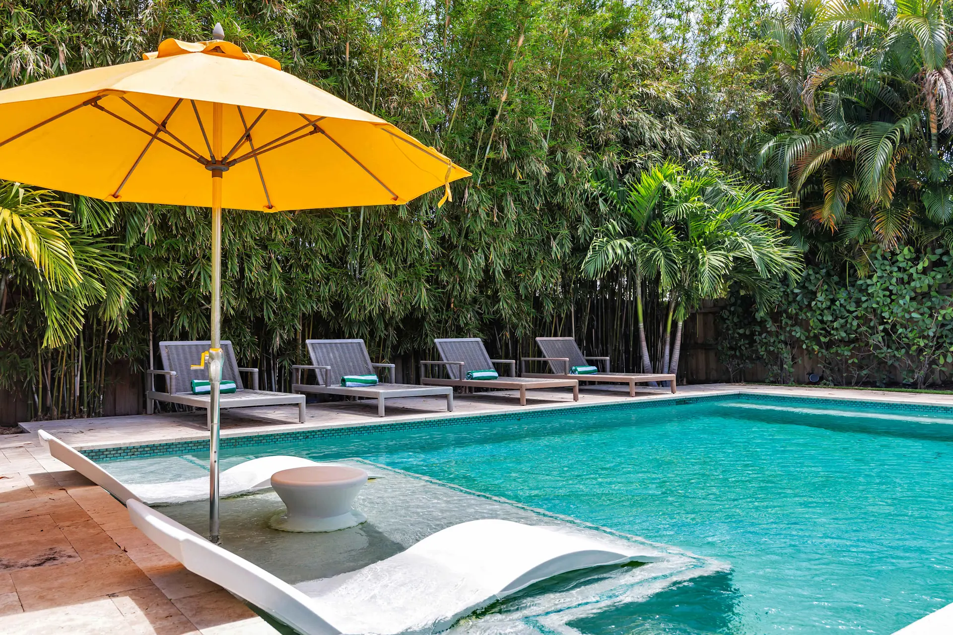 A sun-drenched pool area with lounge chairs, an umbrella, and lush tropical foliage.