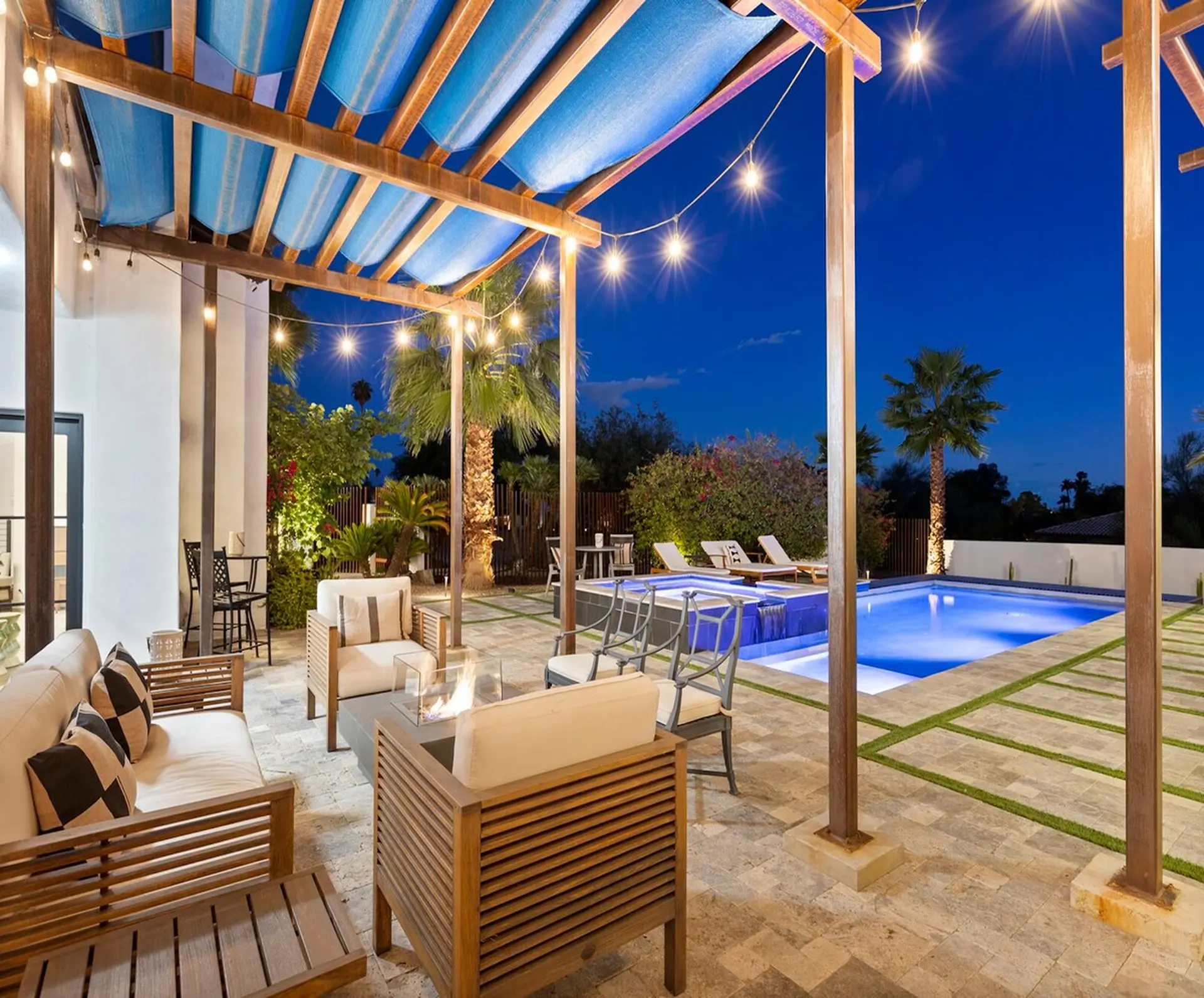 A luxurious patio with a pool, fire pit, and string lights at dusk.
