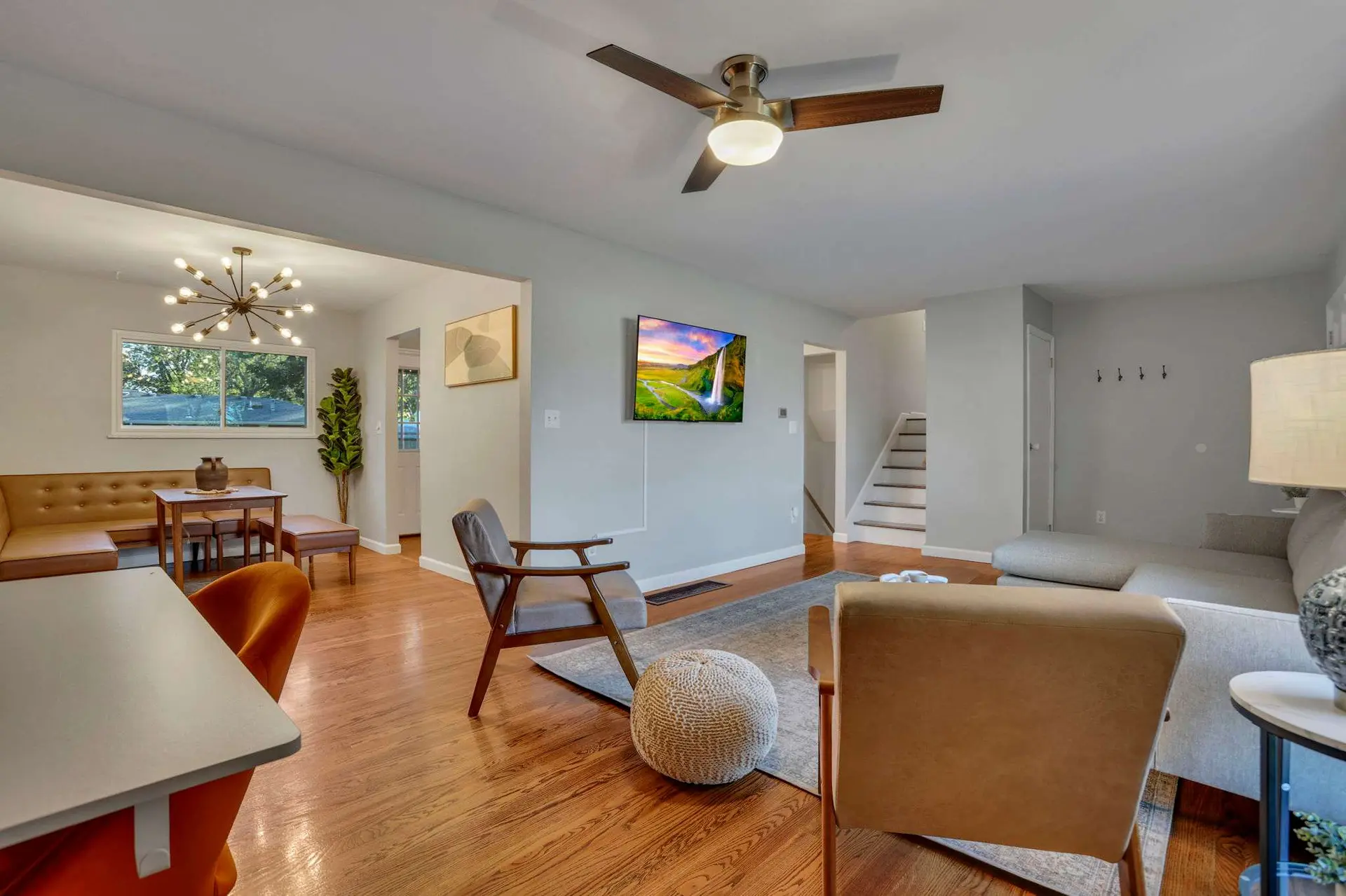 A bright, modern living room with wooden floors, a TV, a ceiling fan, and seating areas.