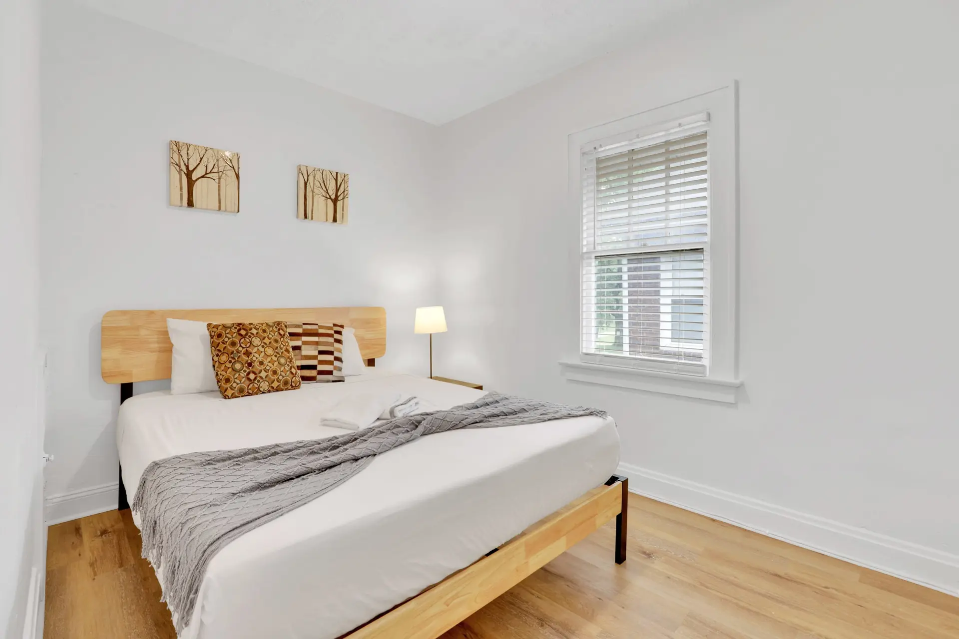 A minimalist bedroom features a light wood bed with white linens, grey blanket, and patterned pillow